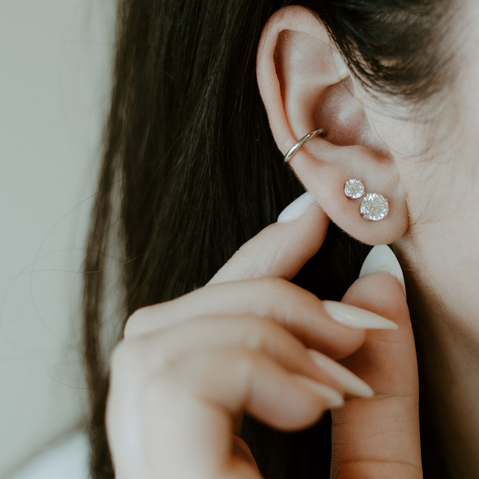 A person's ear with two diamond earrings and a silver ear cuff. Hand touching ear.