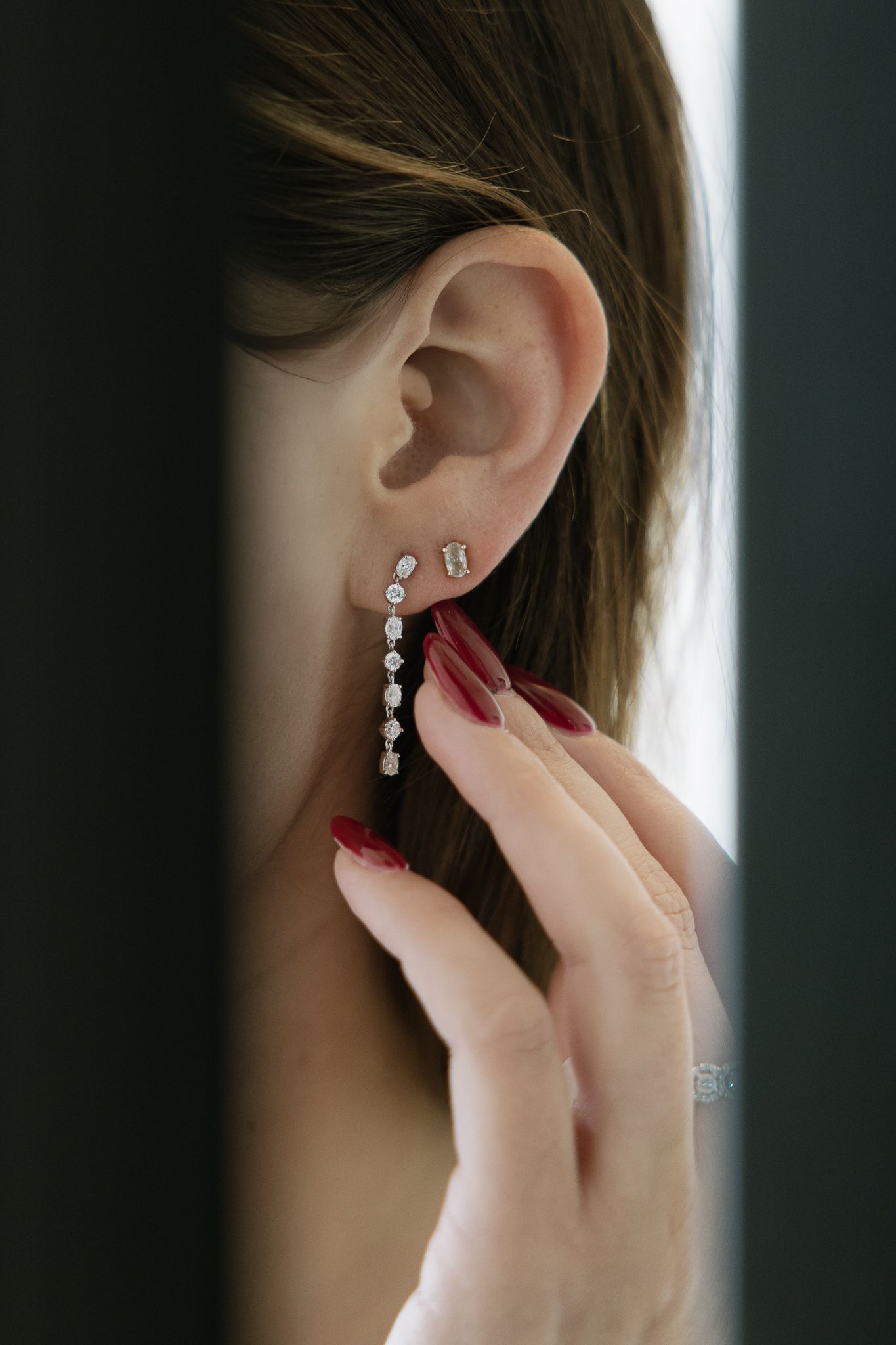 Woman's ear with dangling diamond earrings, hand touching the earring; red nails.