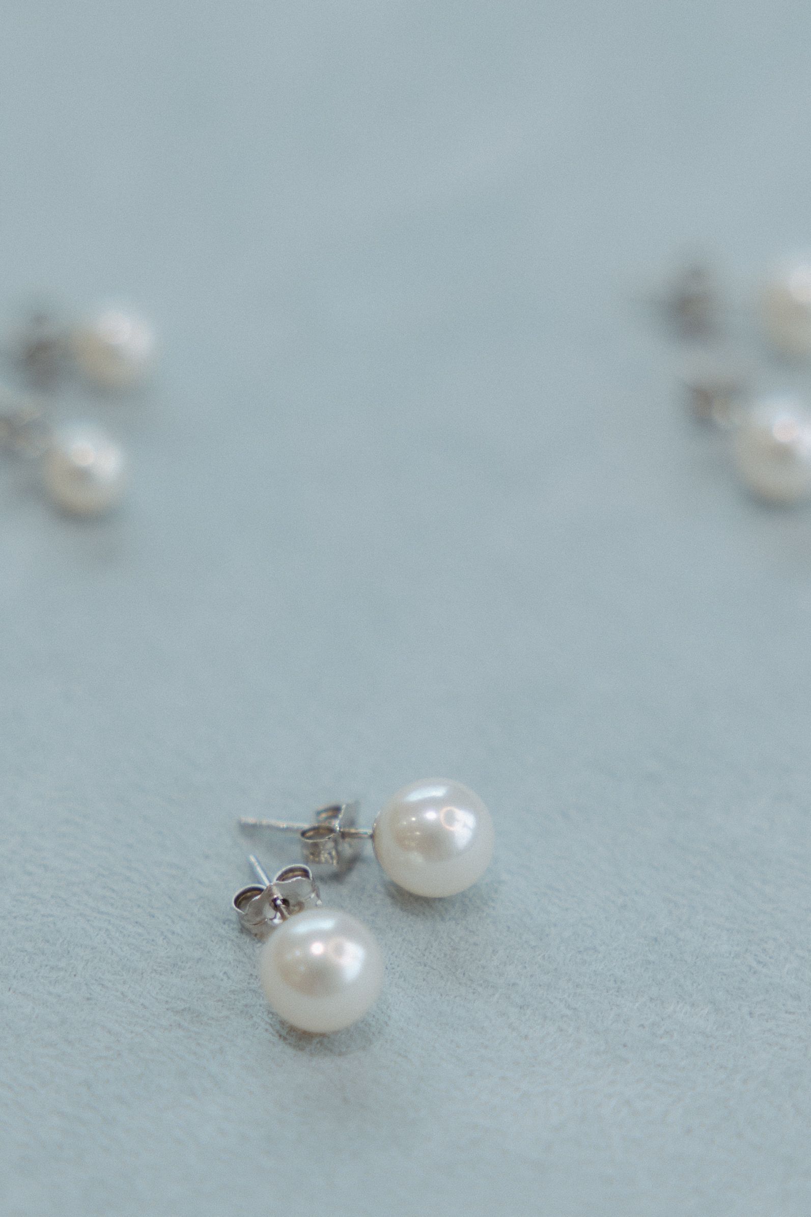 Pair of pearl stud earrings on a light blue surface, with other blurry earrings in background.