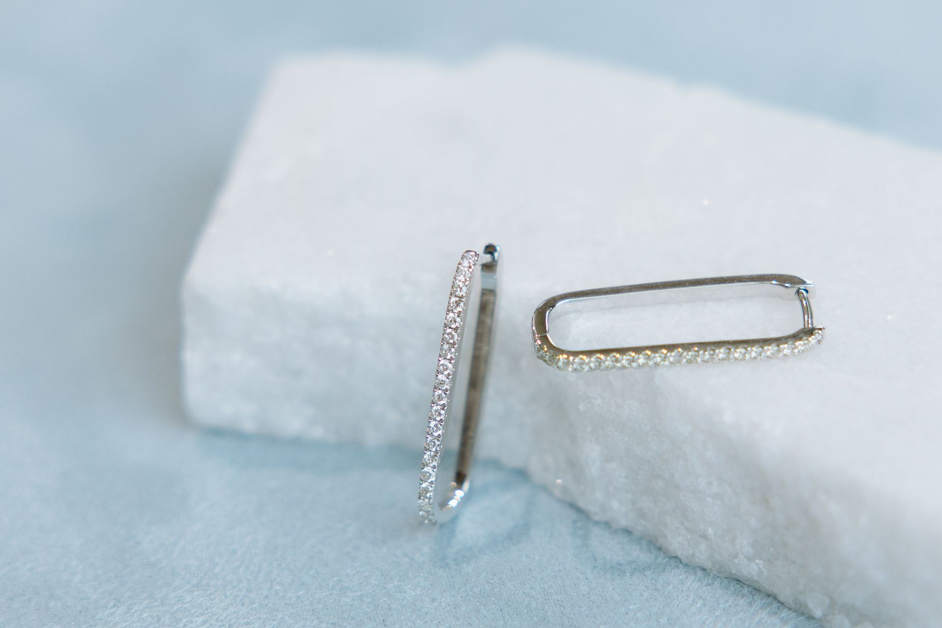 Two rectangular silver earrings with small crystals, resting on a white block against a blue backdrop.