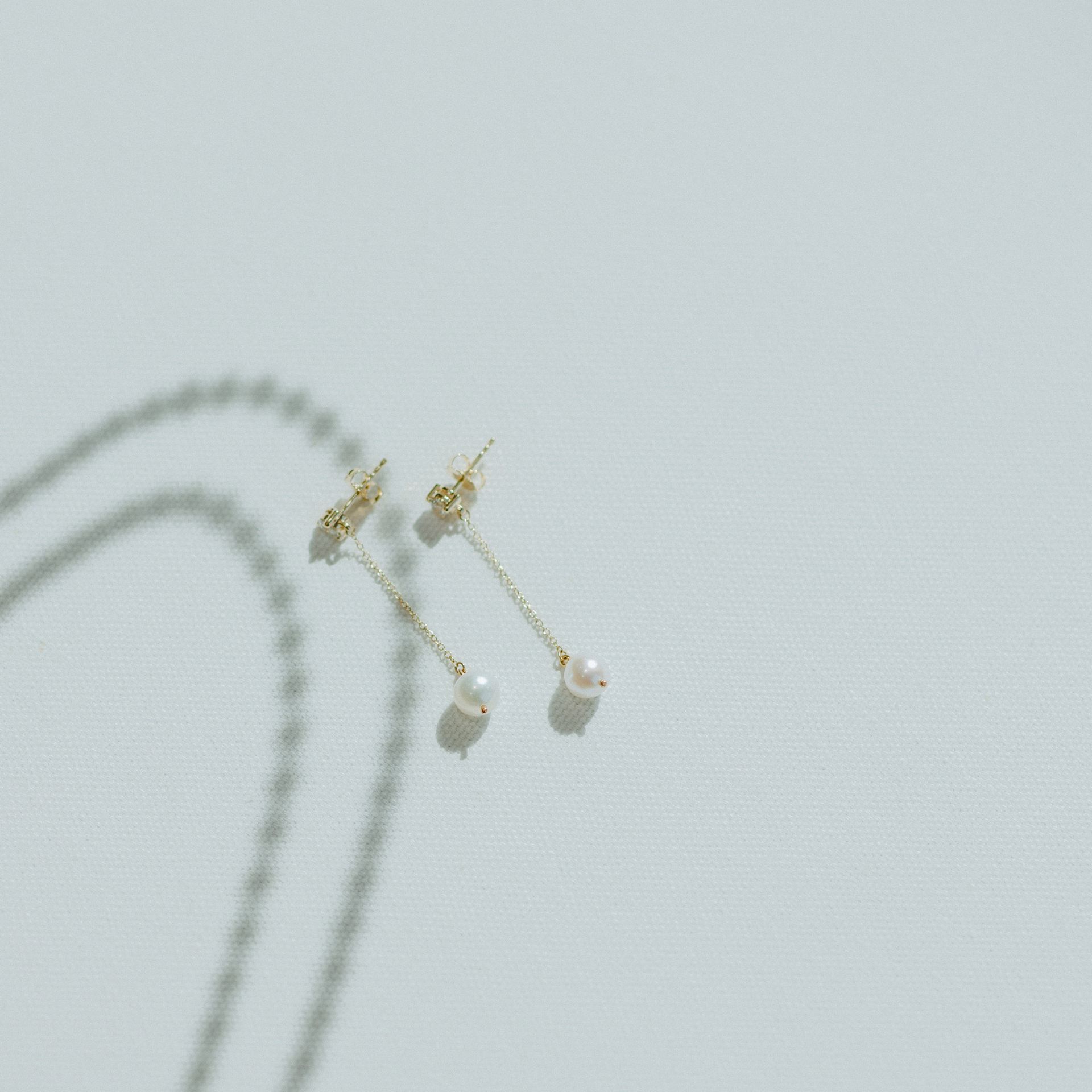 Gold drop earrings with white pearls, lying on a white surface. A chain shadow is in the background.