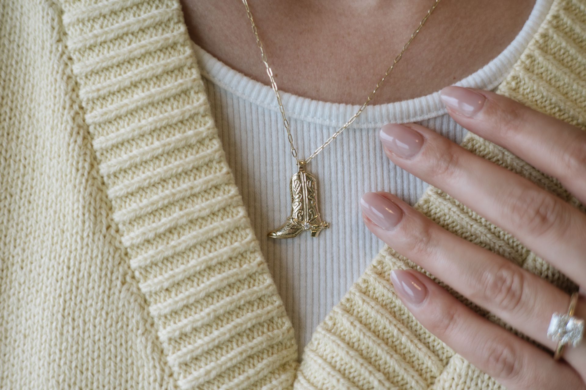 Woman wearing gold necklace with bell pendant, over a white ribbed shirt and yellow sweater. Hand on chest.
