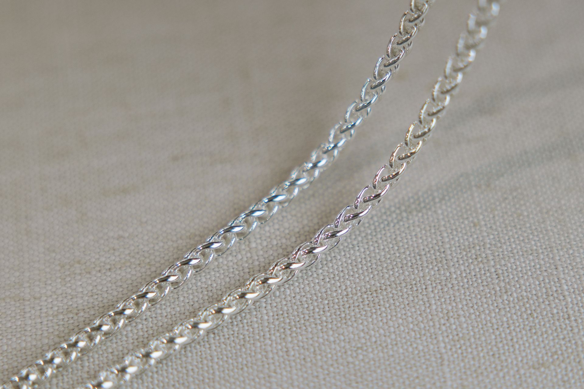 Two silver wheat chain necklaces on a light-colored, textured surface.