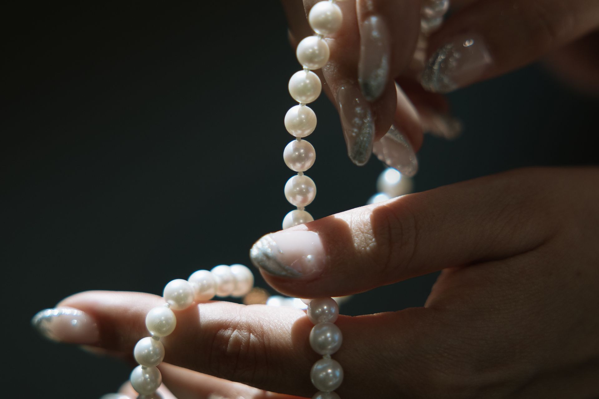 Hands holding a strand of white pearls, against a dark background, sunlight.