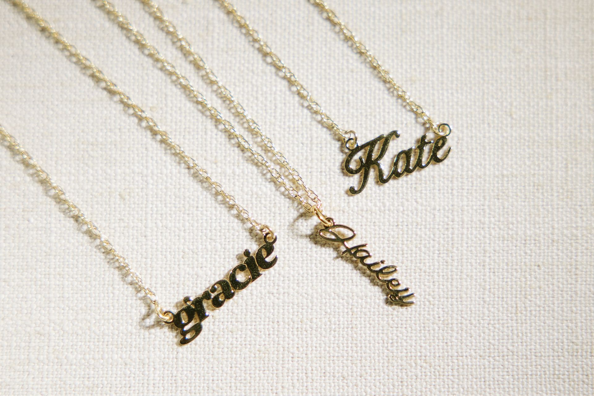Four gold nameplate necklaces on a white surface, spelling Gracie, Kate, and Bailey.