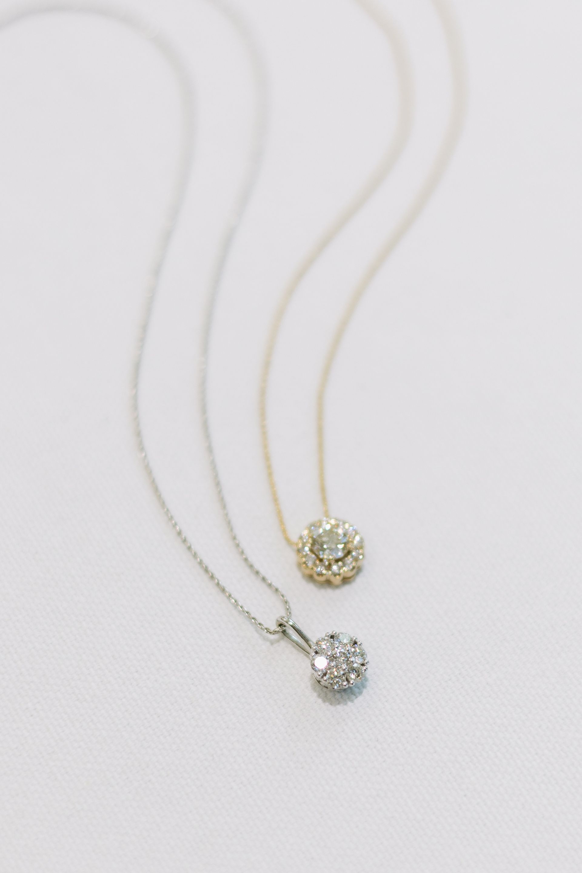 Two diamond pendant necklaces on a white textured surface; one silver, one gold.