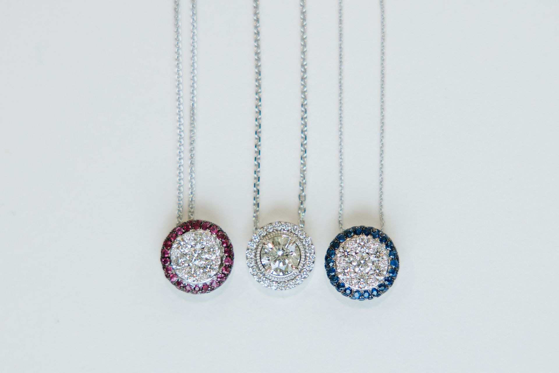 Three silver necklaces with circular pendants: red, clear, and blue gemstone halos around a central cluster.
