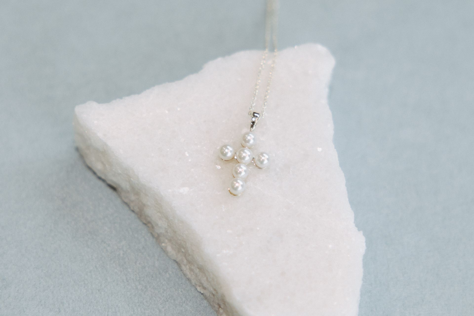 Pearl cross pendant on a white, textured rock against a soft blue background.