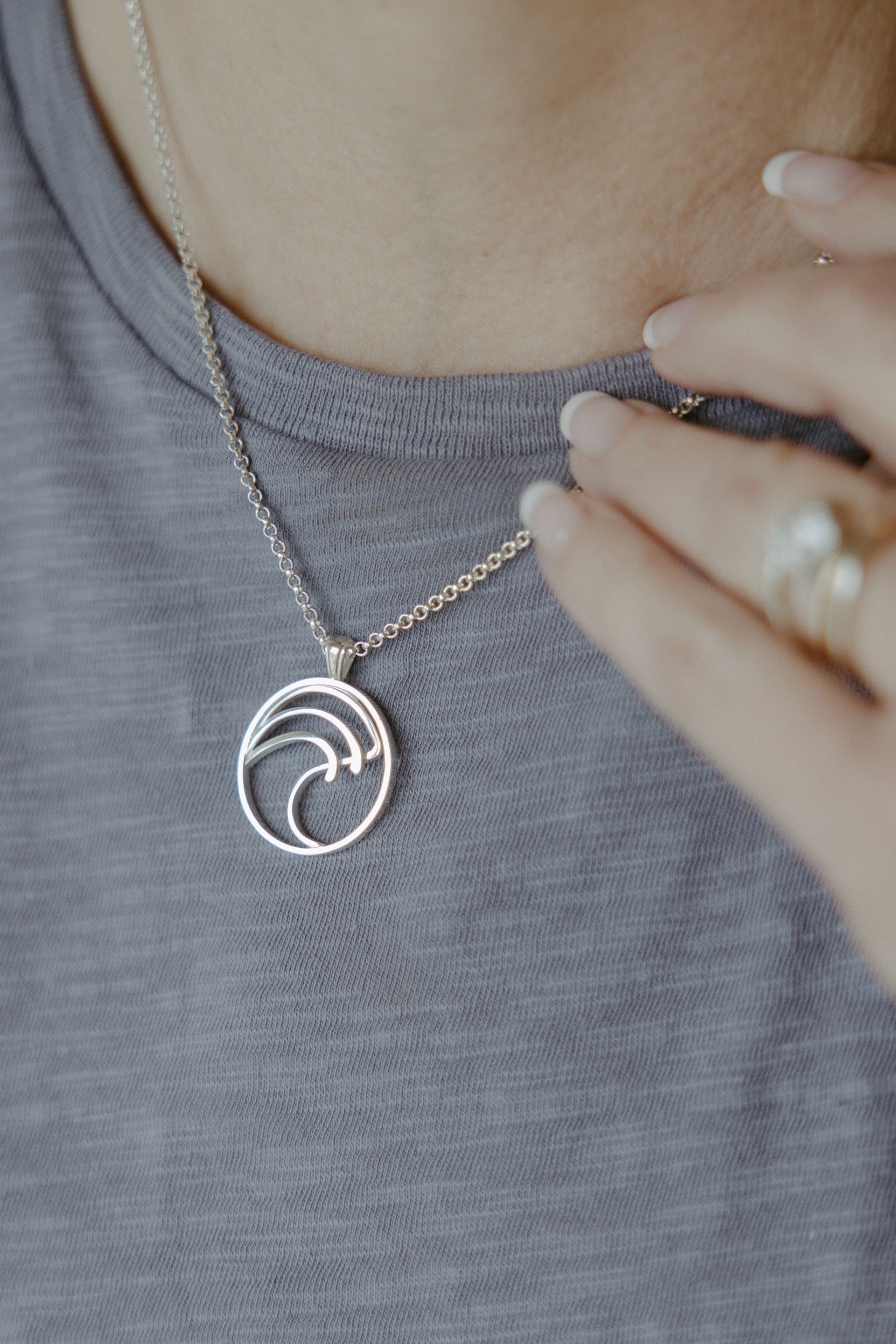 Silver wave pendant necklace on a person wearing a gray shirt, hand touching the collar.