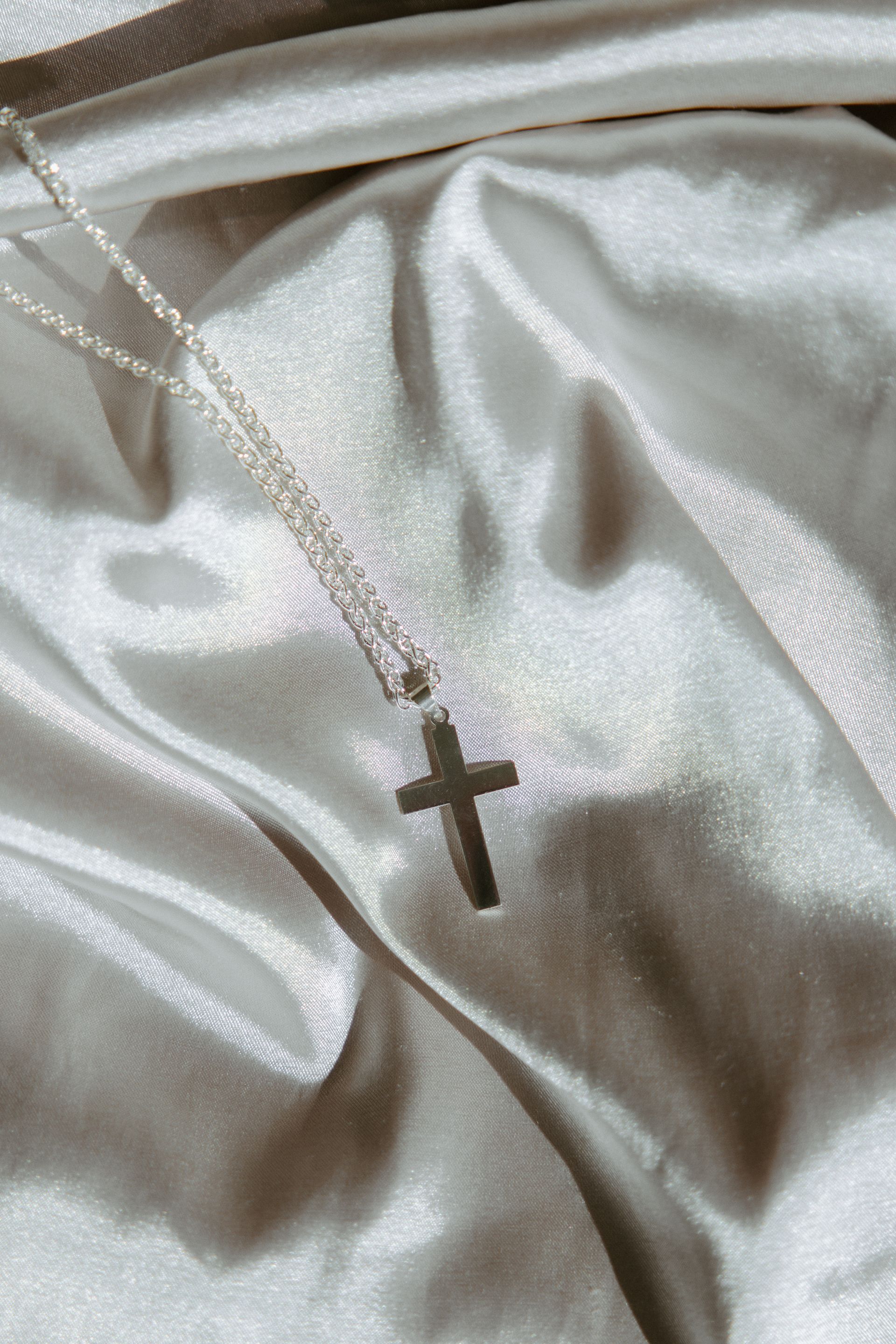 Silver cross pendant on a delicate chain, resting on silvery, draped fabric.