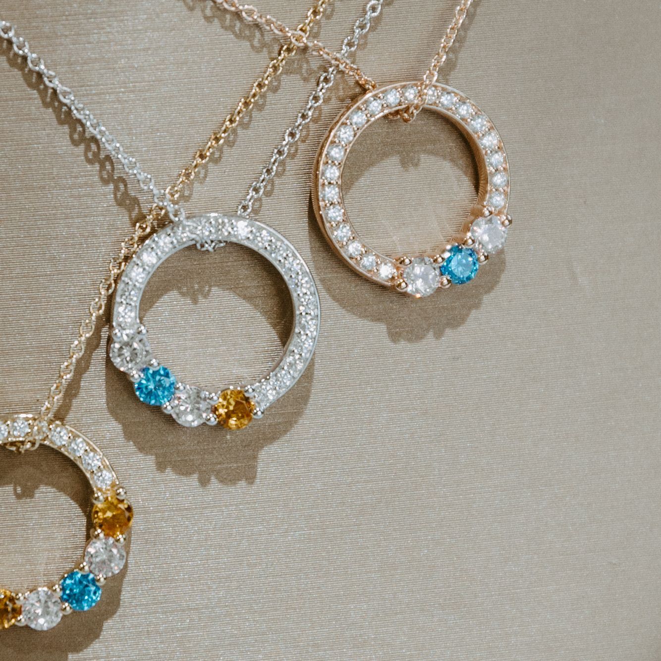 Three diamond circle necklaces with colored gems on a beige surface.