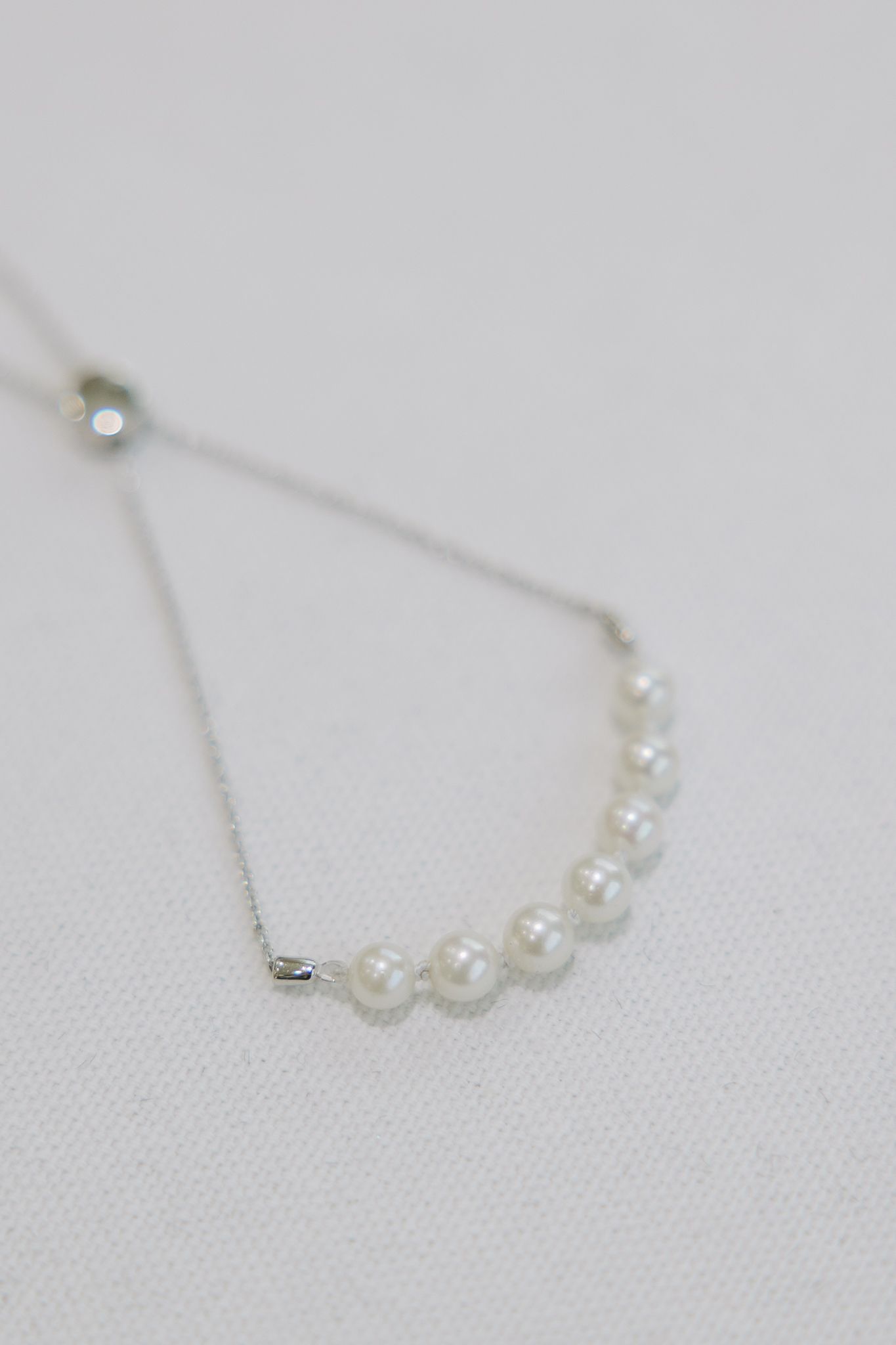 Silver bracelet with a row of small, white pearls, set against a white fabric background.