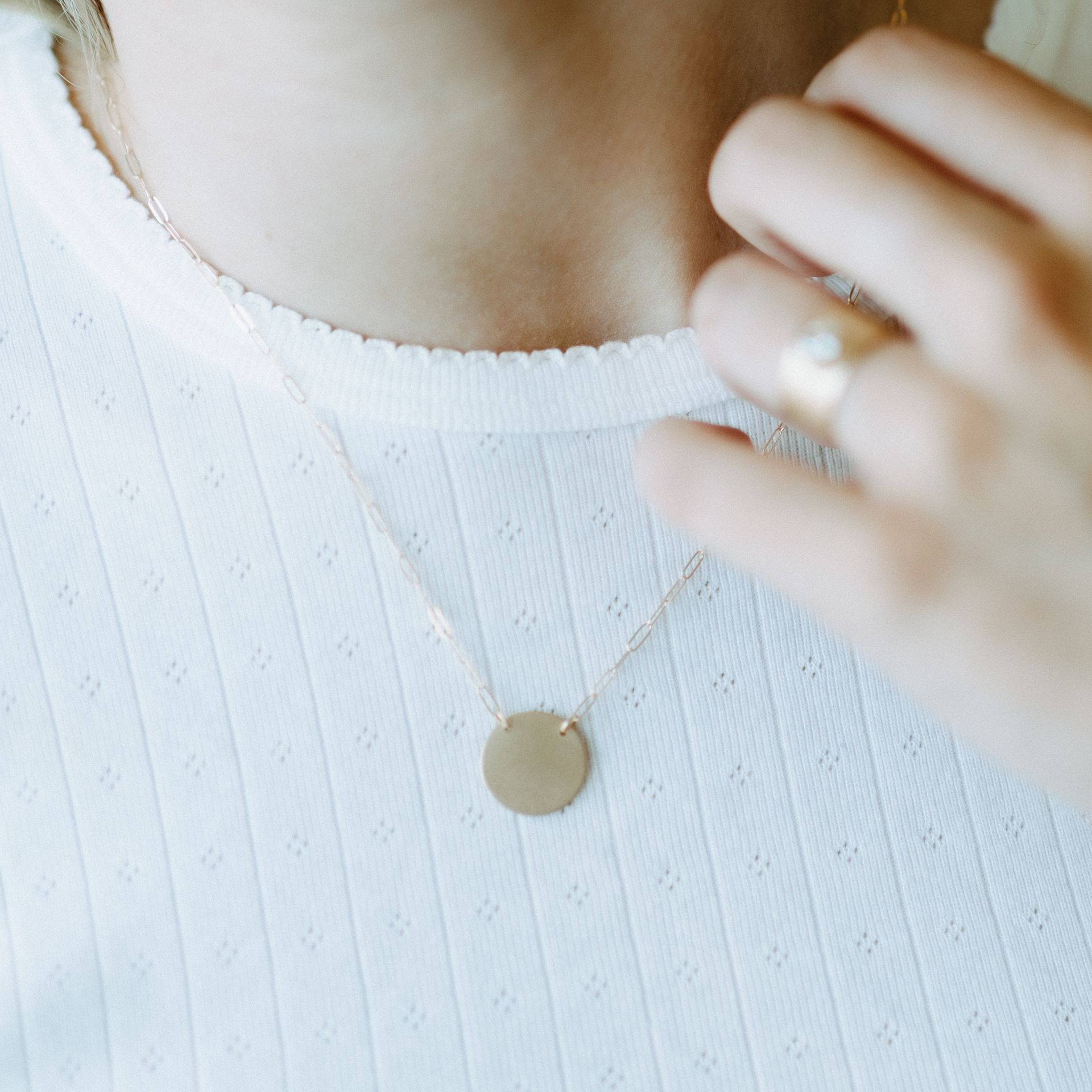 Woman wearing gold pendant necklace, hand touching necklace near the chest, white shirt.