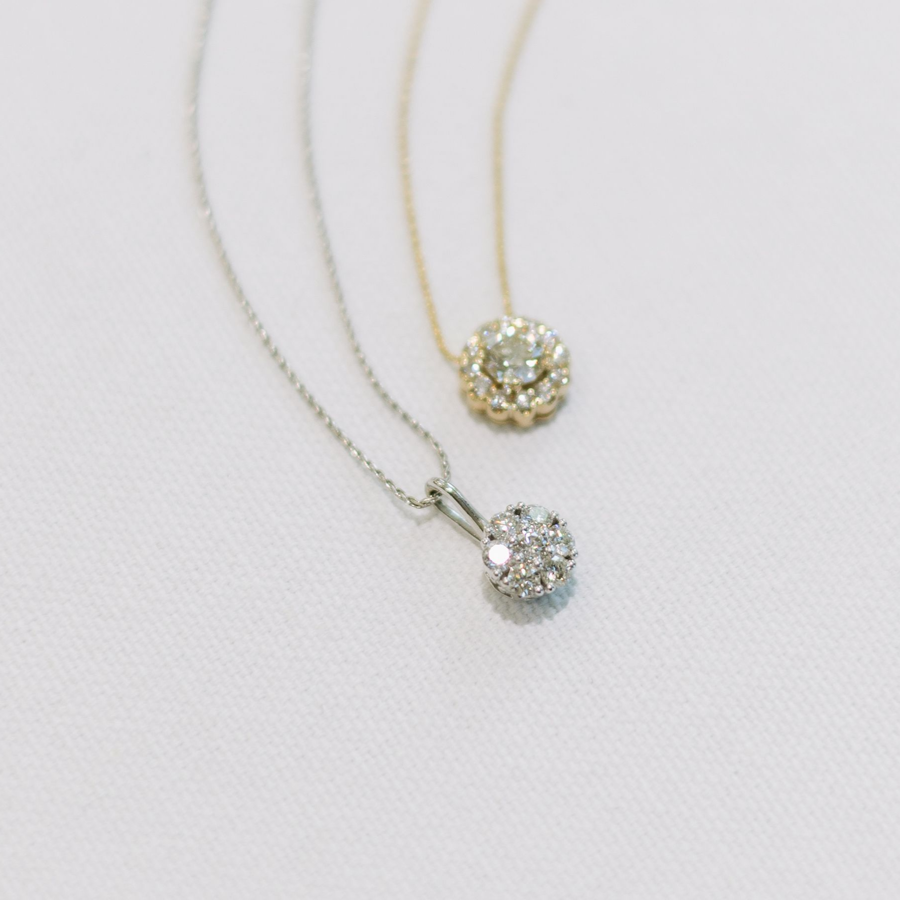 Two diamond pendant necklaces on a white textured surface; one silver, one gold.
