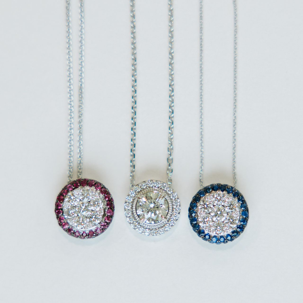 Three silver necklaces with circular pendants: red, clear, and blue gemstone halos around a central cluster.