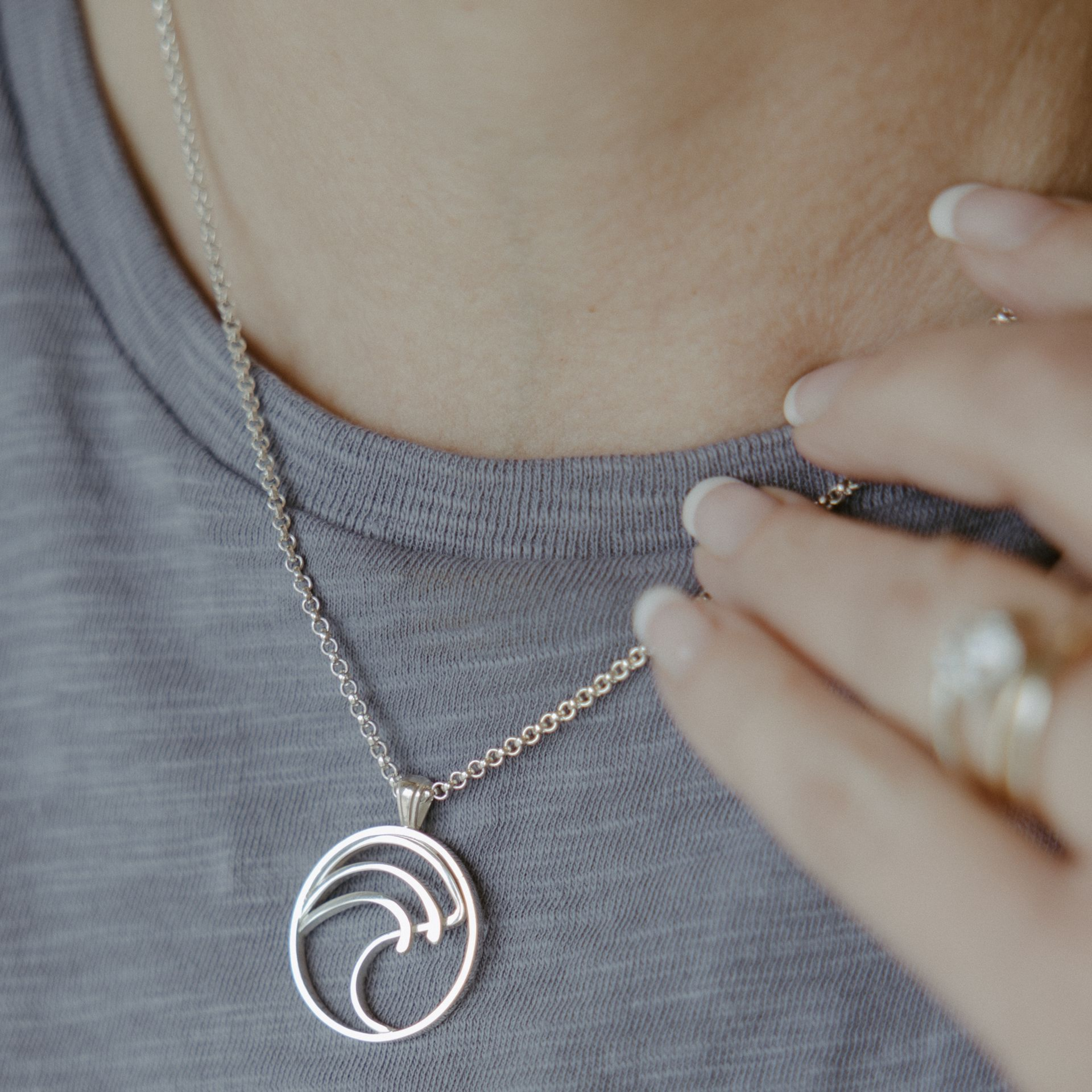 Silver wave pendant necklace on a person wearing a gray shirt, hand touching the collar.