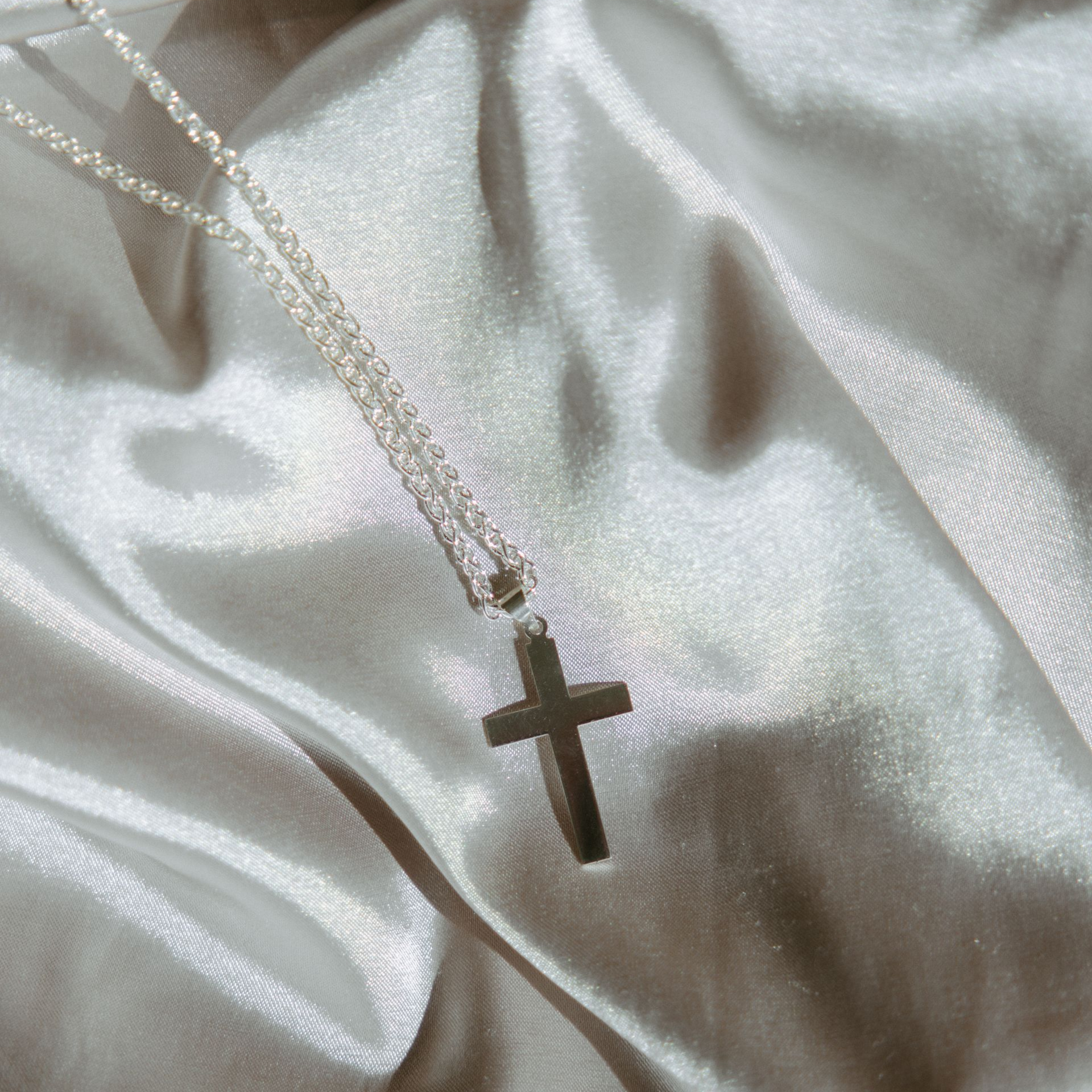 Silver cross pendant on a delicate chain, resting on silvery, draped fabric.