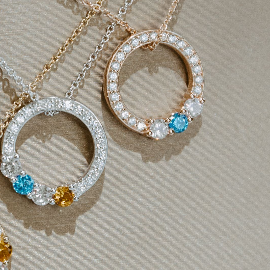 Three diamond circle necklaces with colored gems on a beige surface.