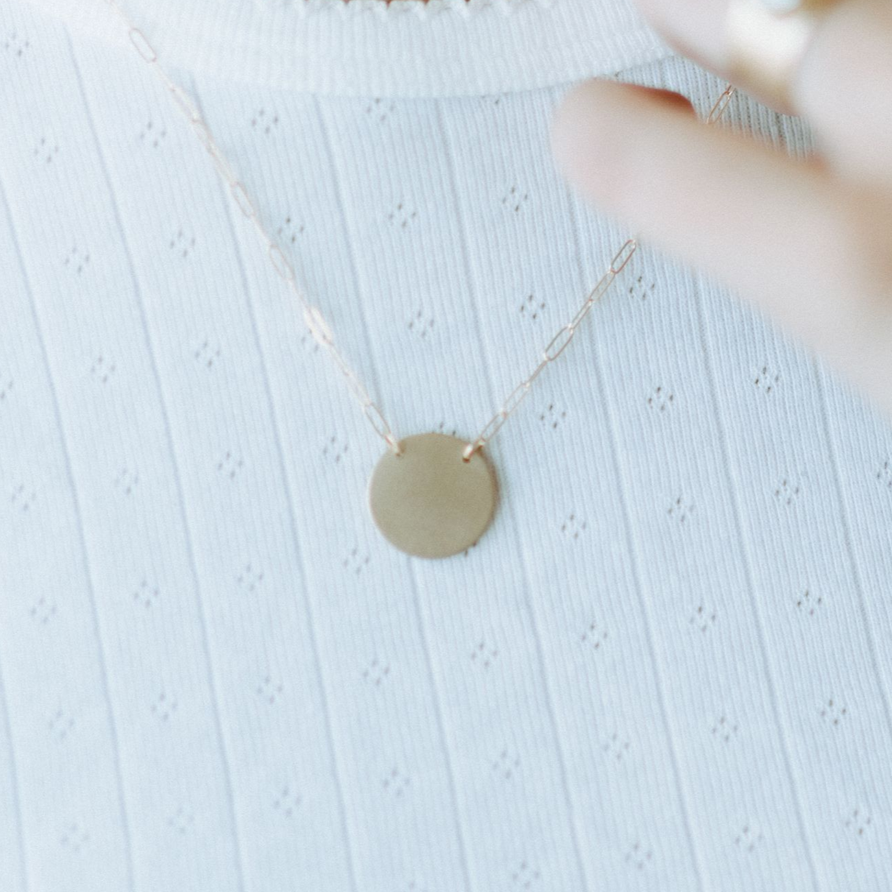 Woman wearing gold pendant necklace, hand touching necklace near the chest, white shirt.