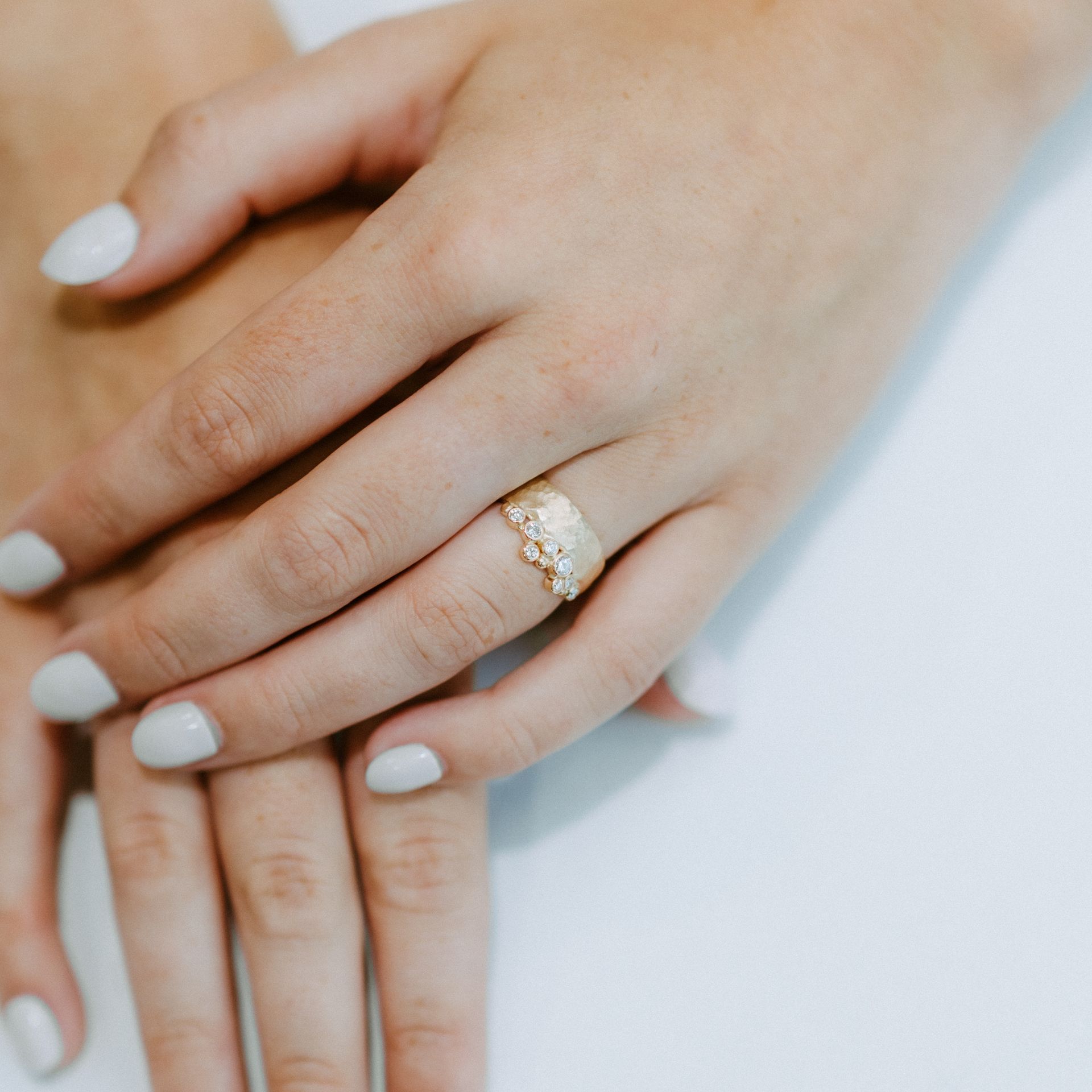 Close-up of hands with light gray nail polish; a gold ring with textured design is on the ring finger.
