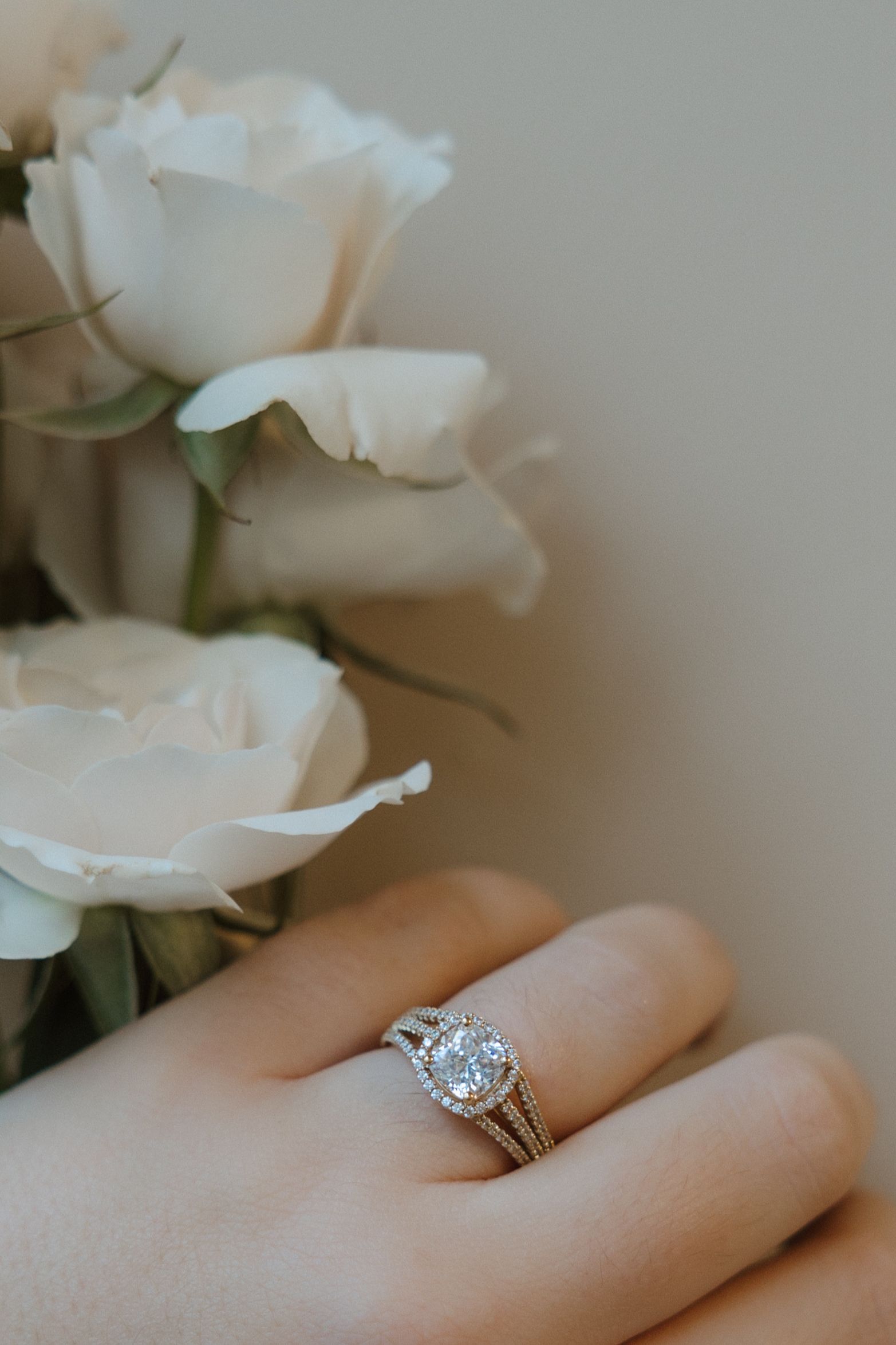 Hand wearing a diamond engagement ring, white roses in the background.