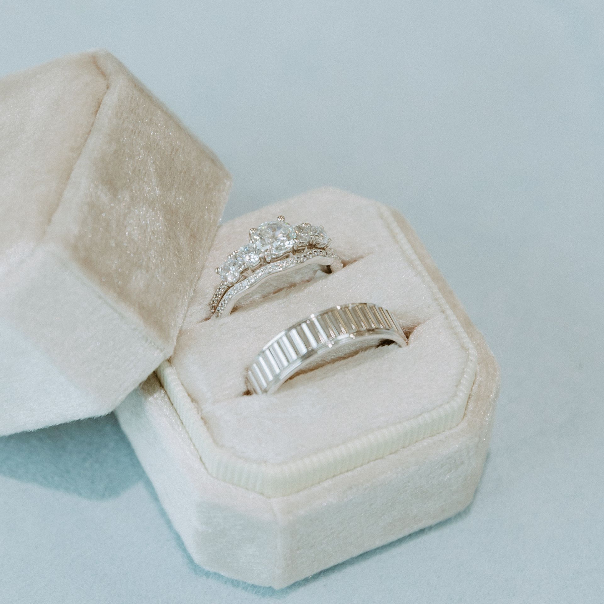 Two wedding rings inside a soft, cream-colored octagonal ring box. One ring has stones, the other is ridged.