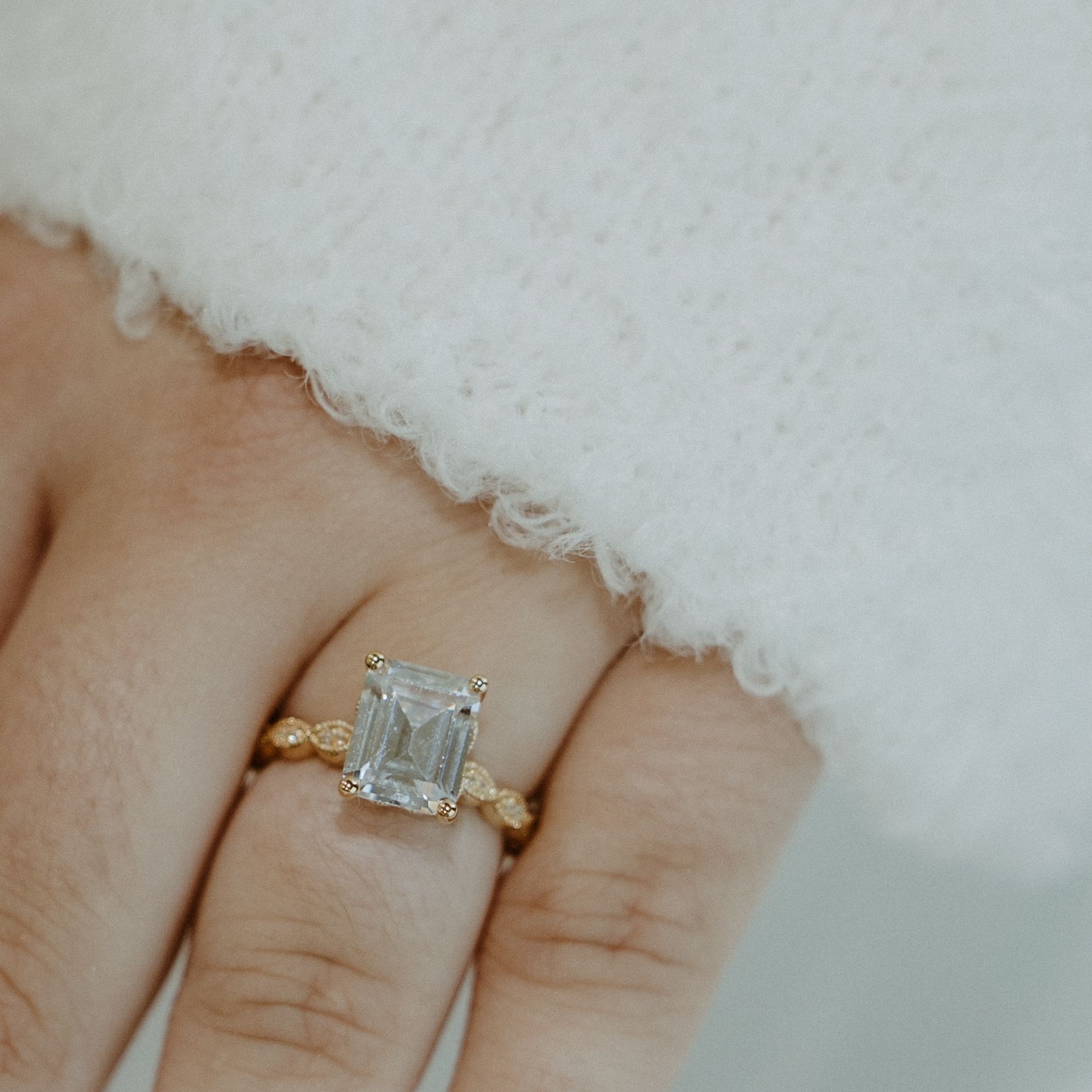 Gold ring with an emerald-cut gemstone on a finger, next to a white sweater.
