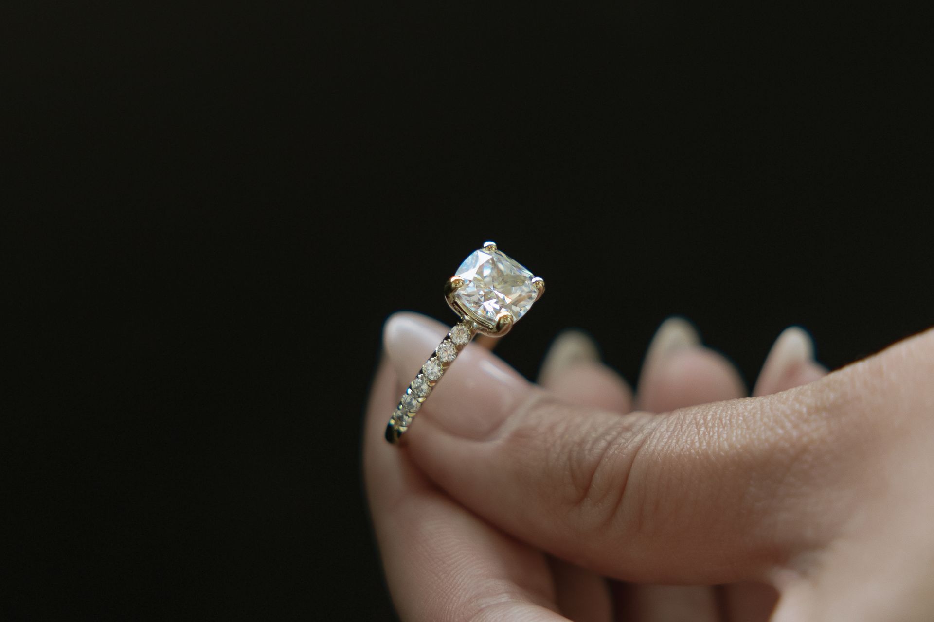 Hand holding a diamond engagement ring with a square-cut stone, against a dark background.
