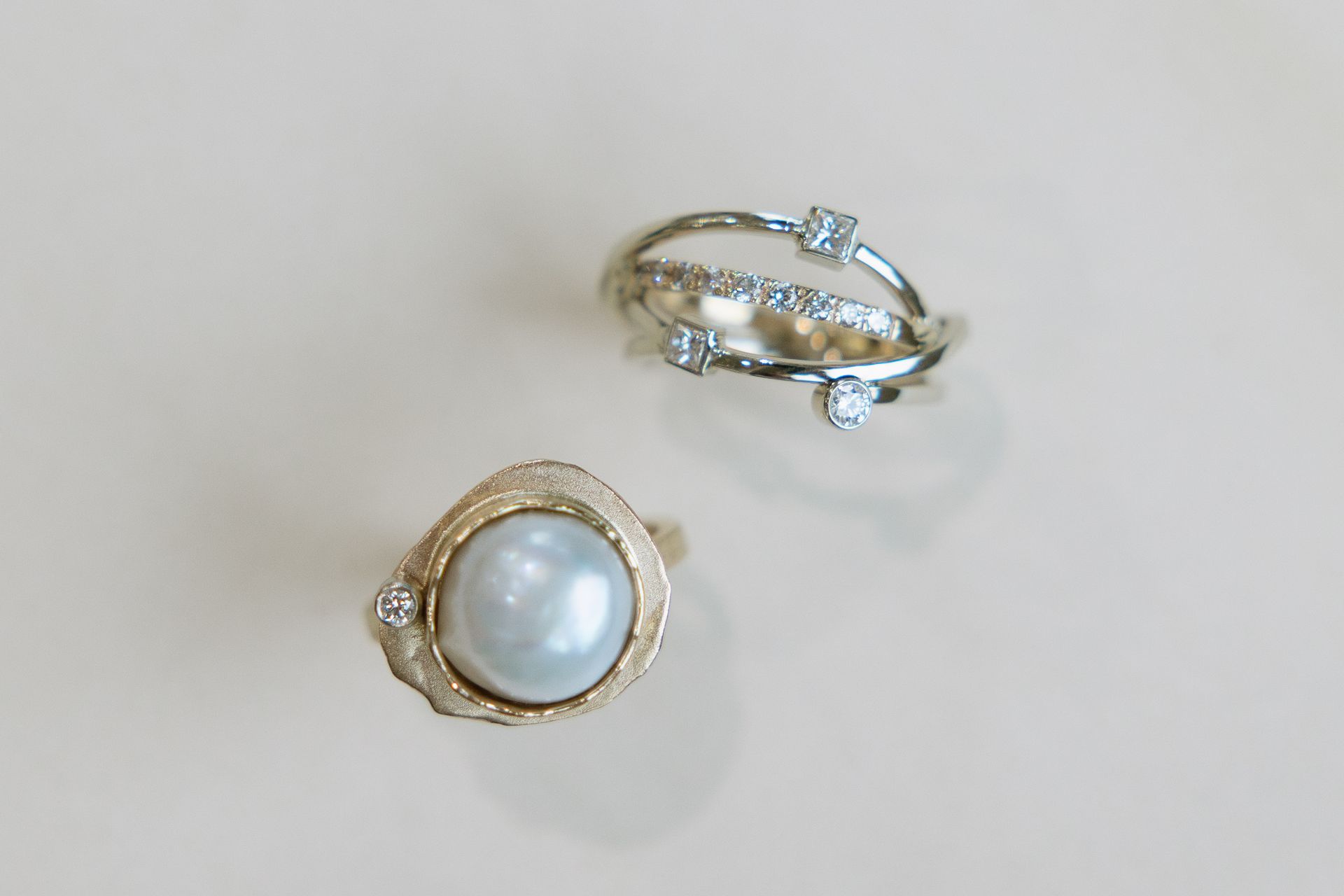 Two ornate rings on a neutral background. One has a large pearl, the other has diamonds and a layered design.