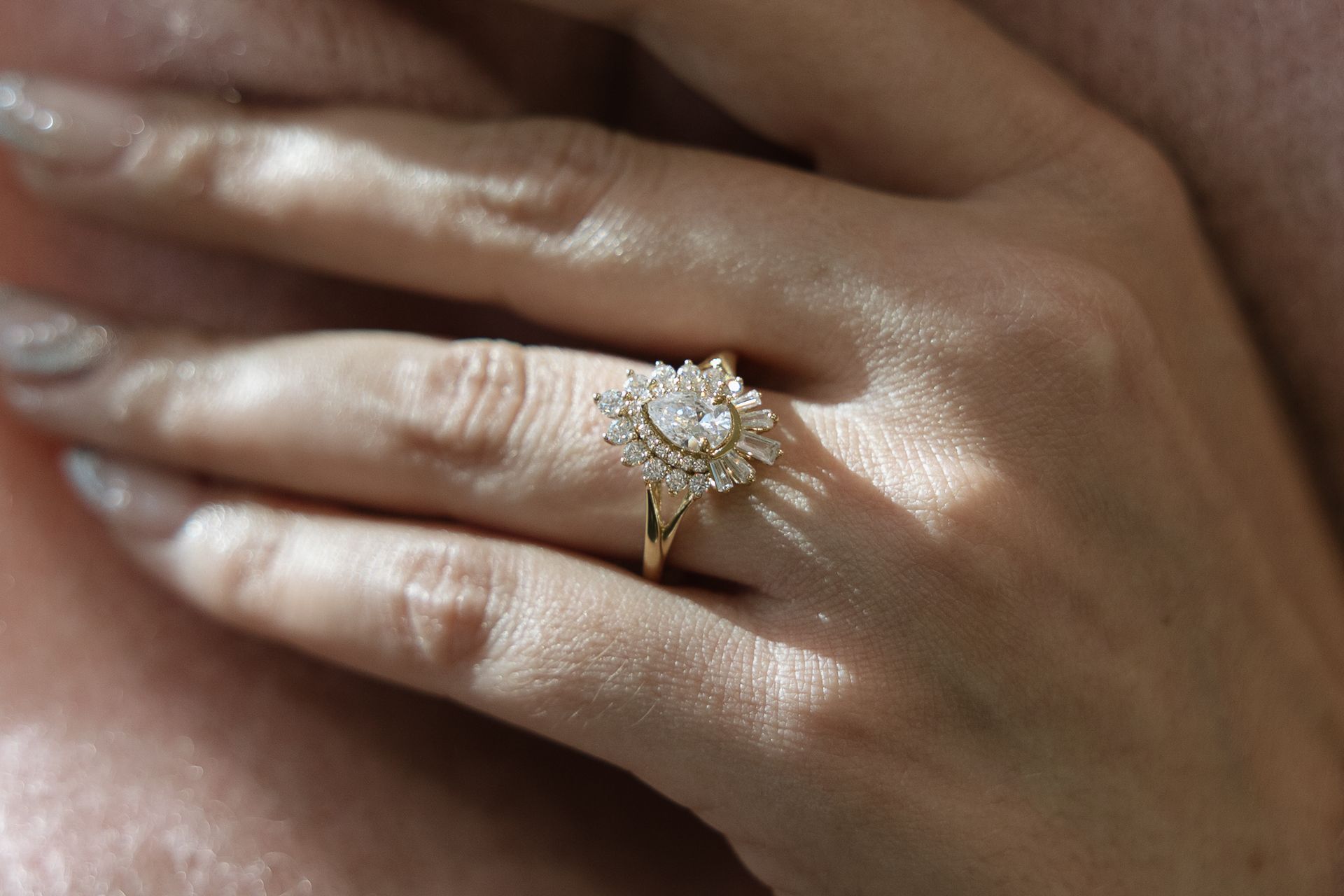 Hand with a gold engagement ring featuring a central pear-shaped stone surrounded by smaller diamonds.