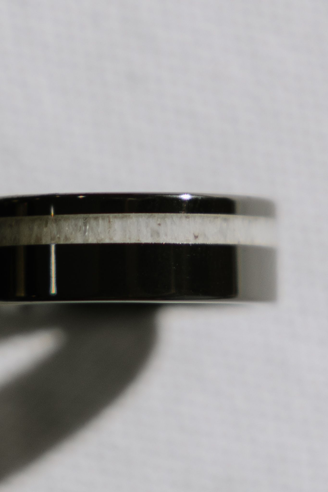 Close-up of a black ring with a light-colored inlay band, set against a blurred background.