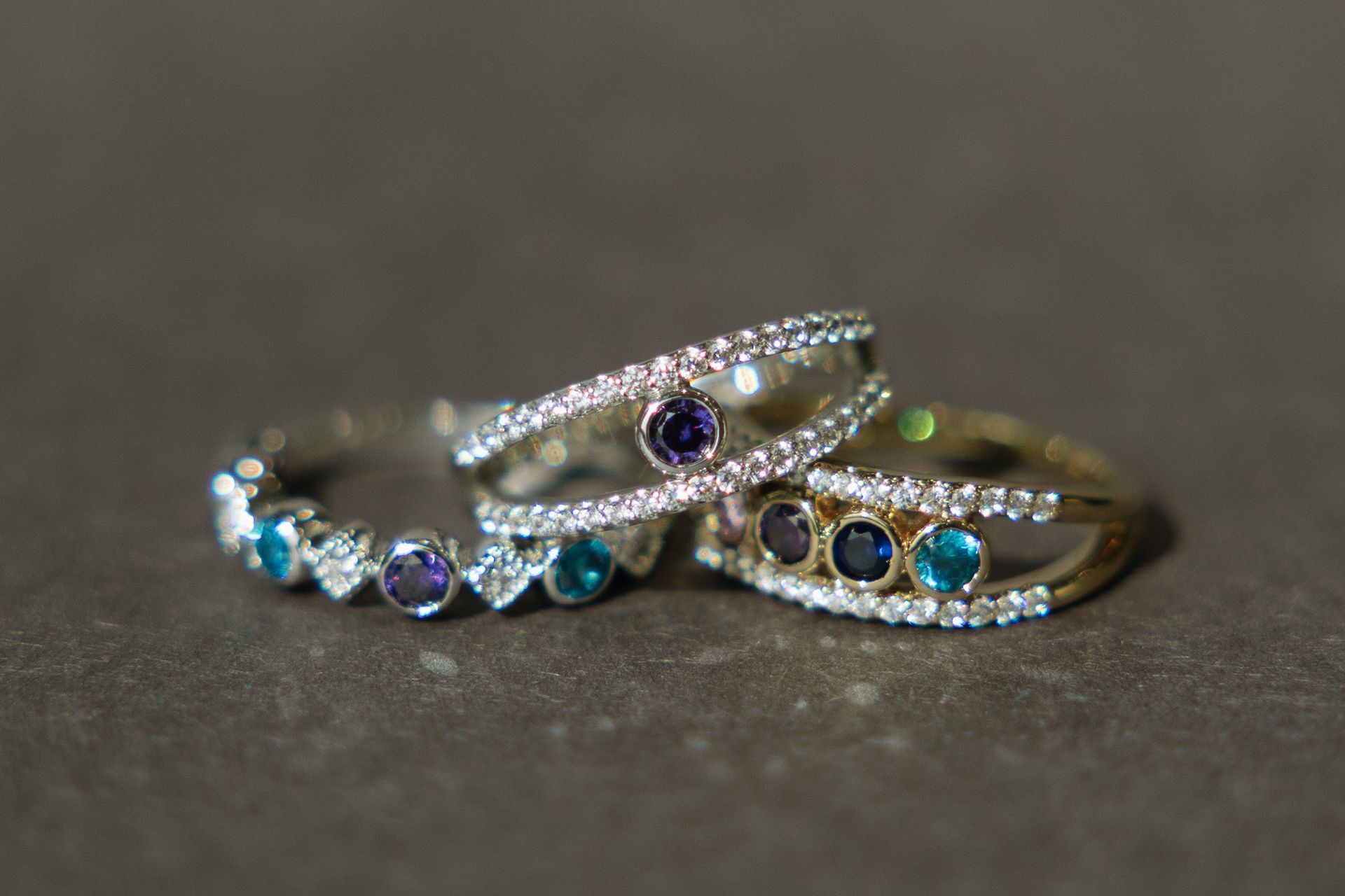 Three gemstone rings: teal, purple, and blue stones in diamond bands, stacked on a gray surface.