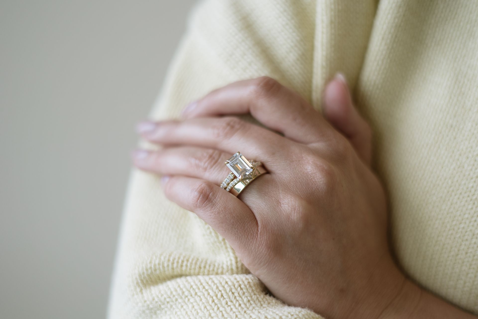 Hand with large emerald-cut diamond ring, gold band, resting on another hand; pale yellow sweater.