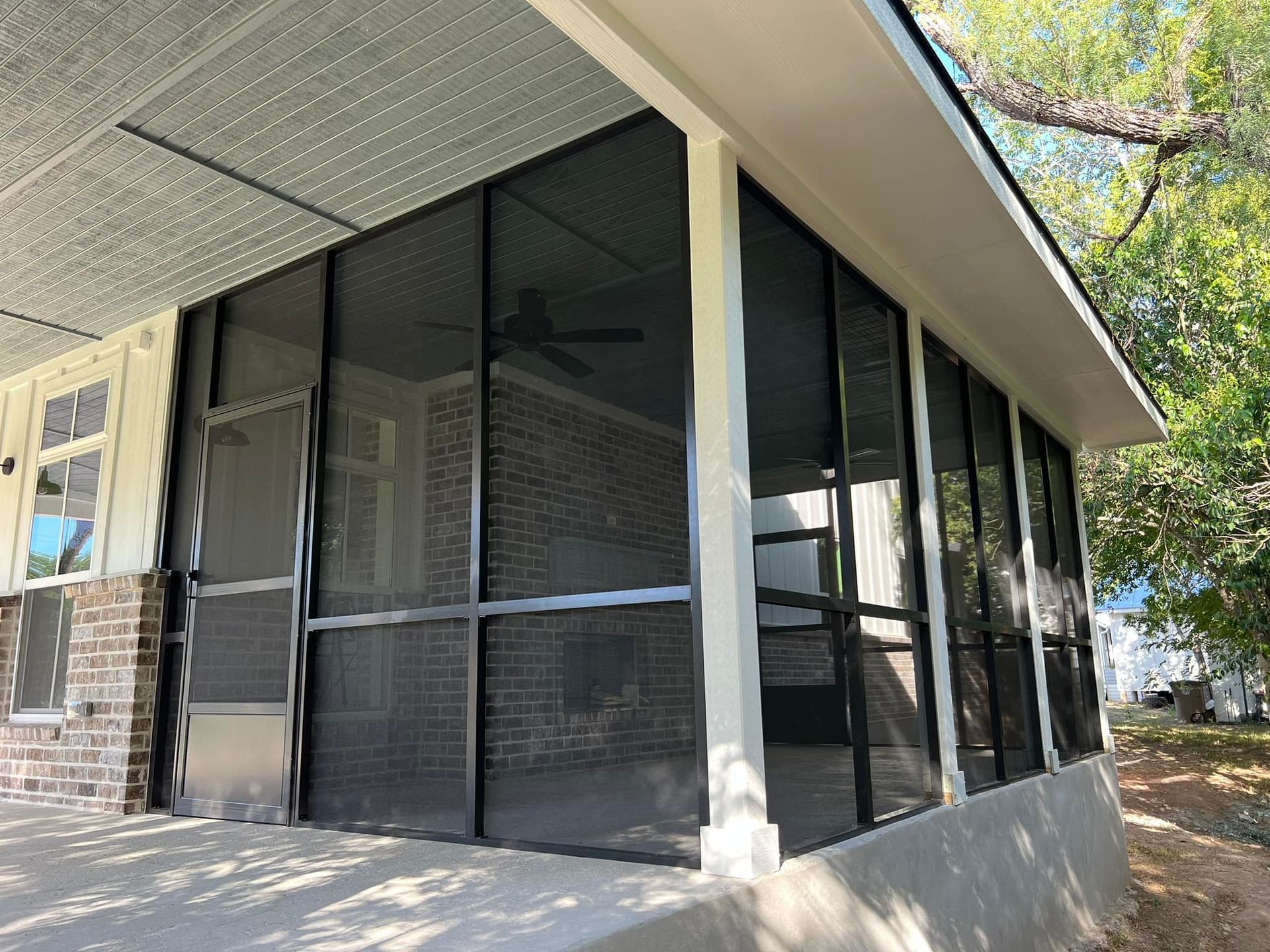A screened in porch with a ceiling fan and a brick house in the background.