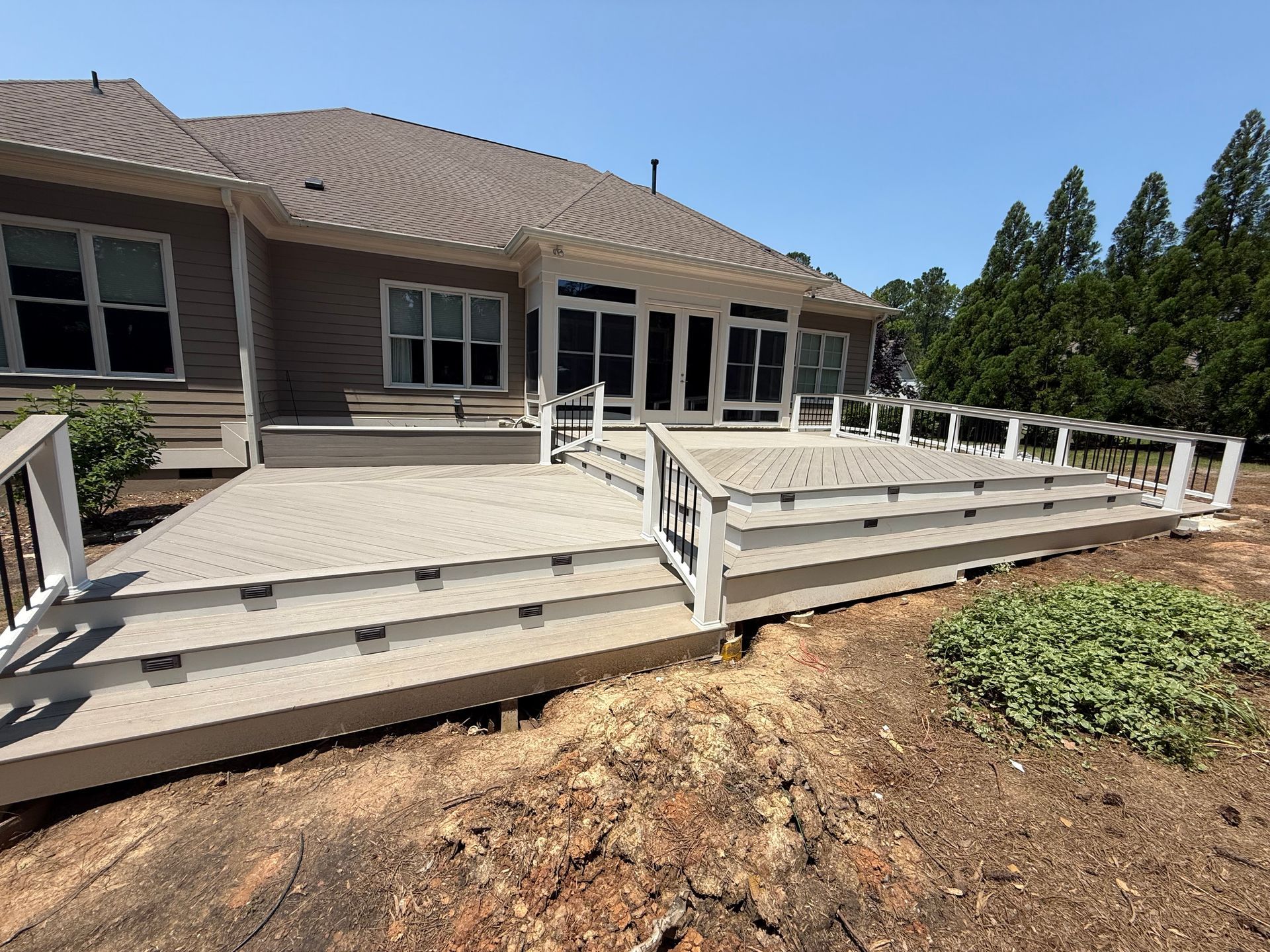 A new composite deck with steps leading to a house, surrounded by dirt and foliage on a sunny day.