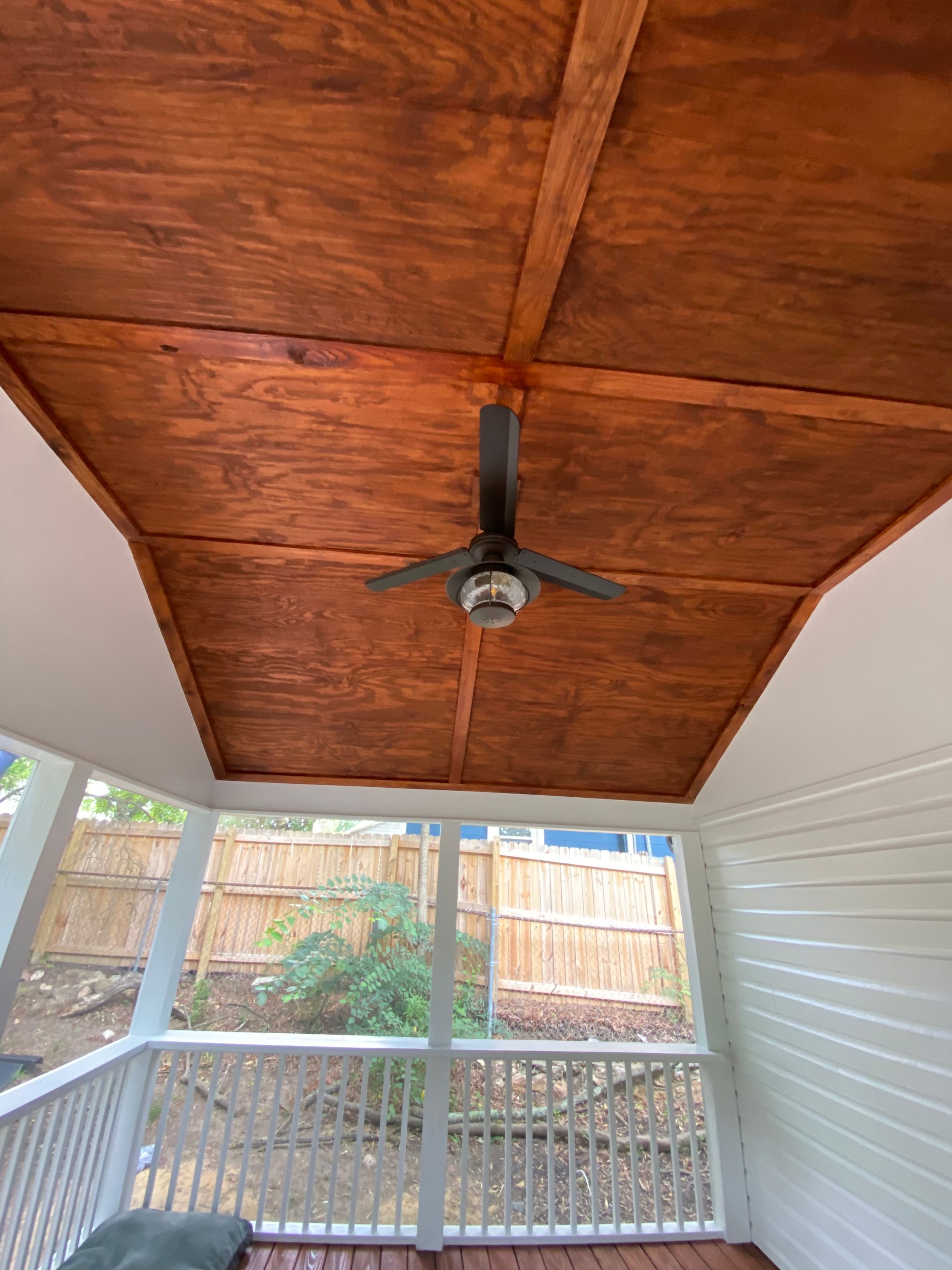Brown wooden ceiling with a black ceiling fan over a porch with a white railing.