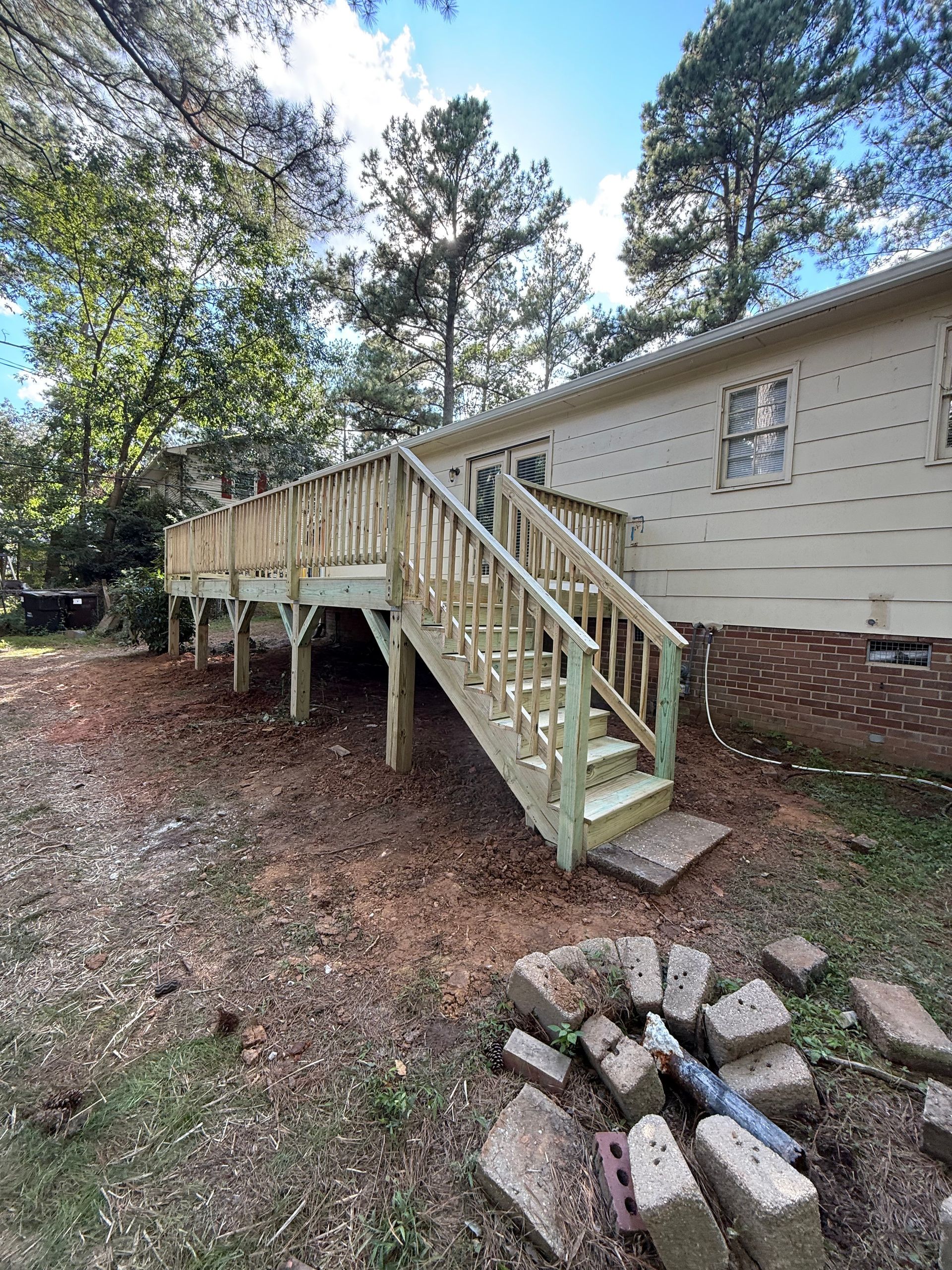 Newly built wooden deck with stairs attached to a light-colored house. Green trees and sky visible.