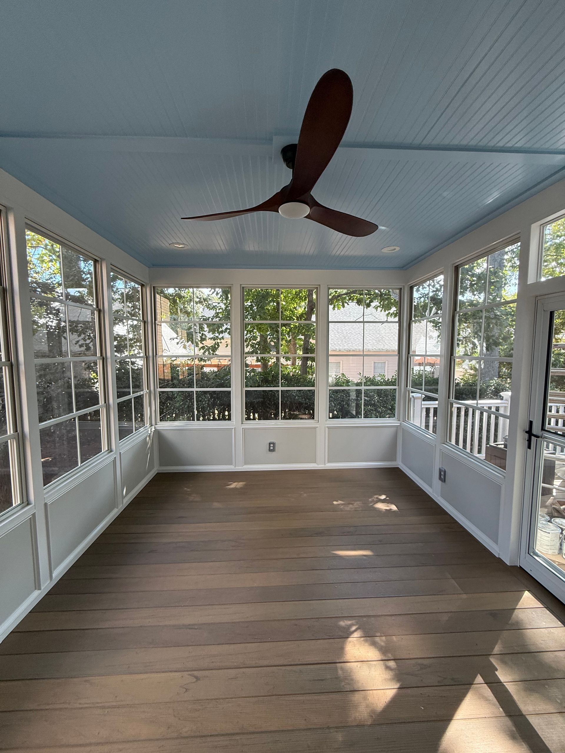 Sunroom with blue ceiling, white trim, and brown wooden floor. Large windows with a ceiling fan.