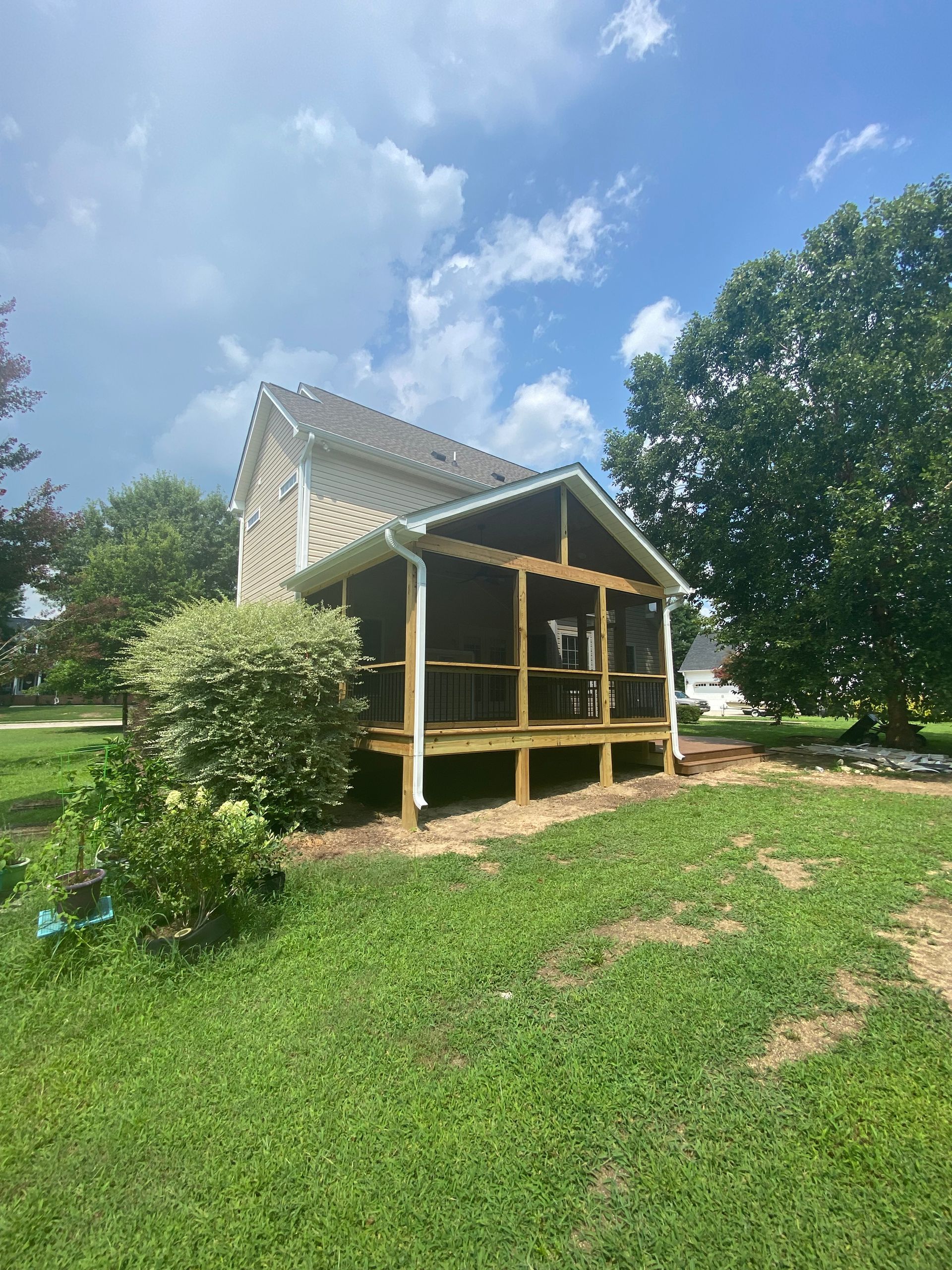 Screened porch attached to a light-colored house. Green grass and trees surround it under a blue sky.