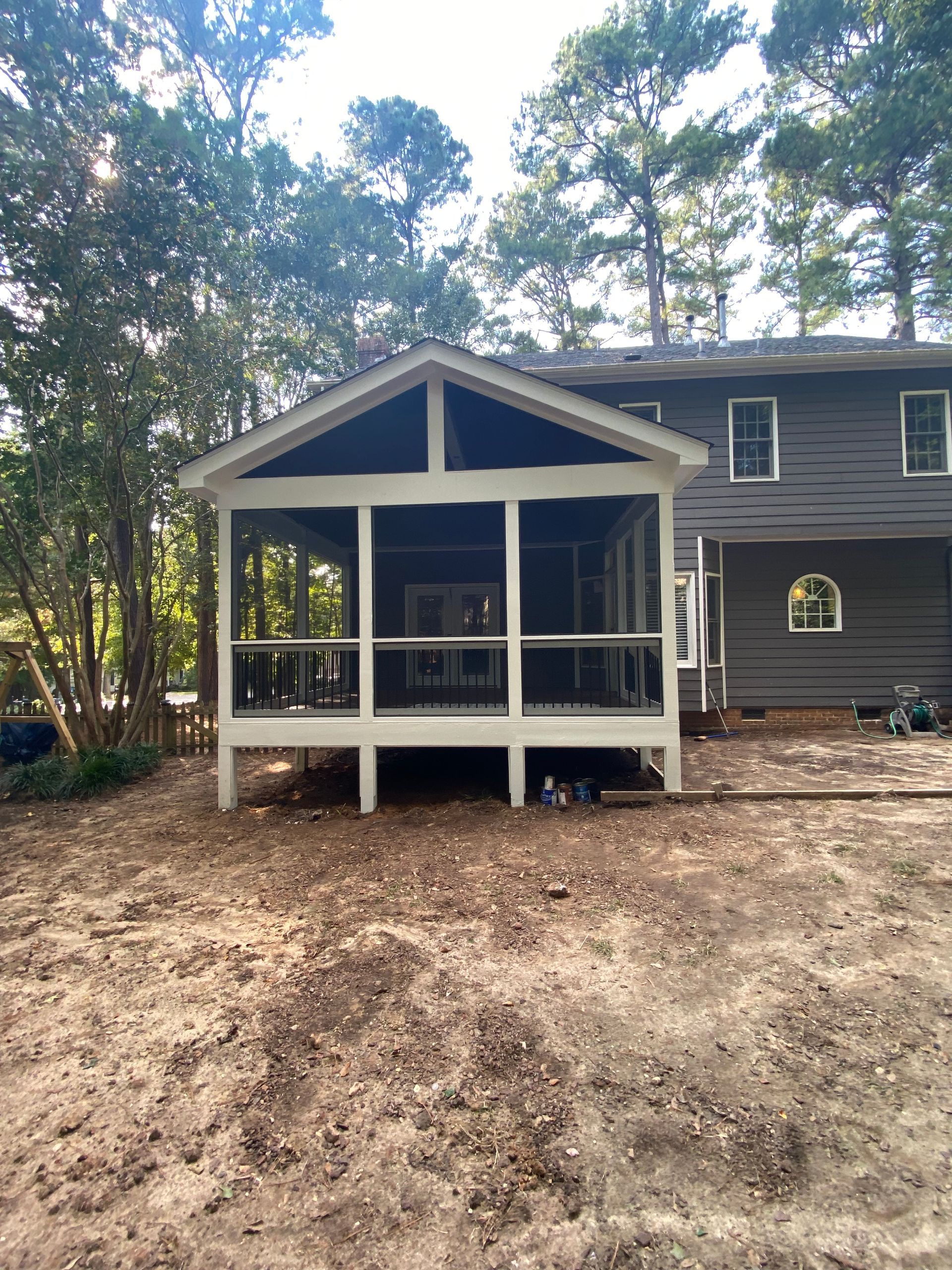 Screened porch with white supports attached to a gray house; trees in the background.