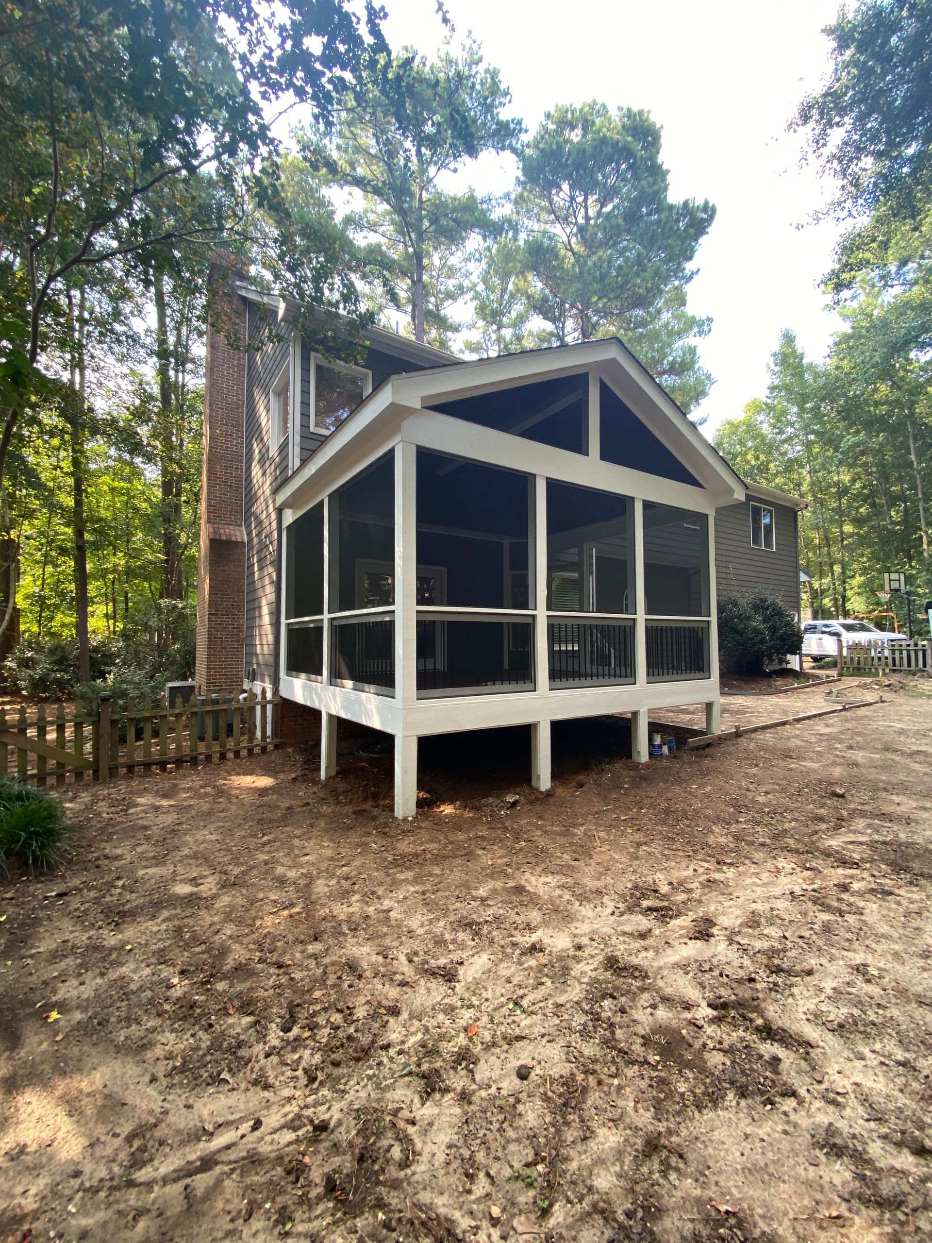Screened-in porch attached to a house; white frame, dark screen, brown backyard, trees in background.