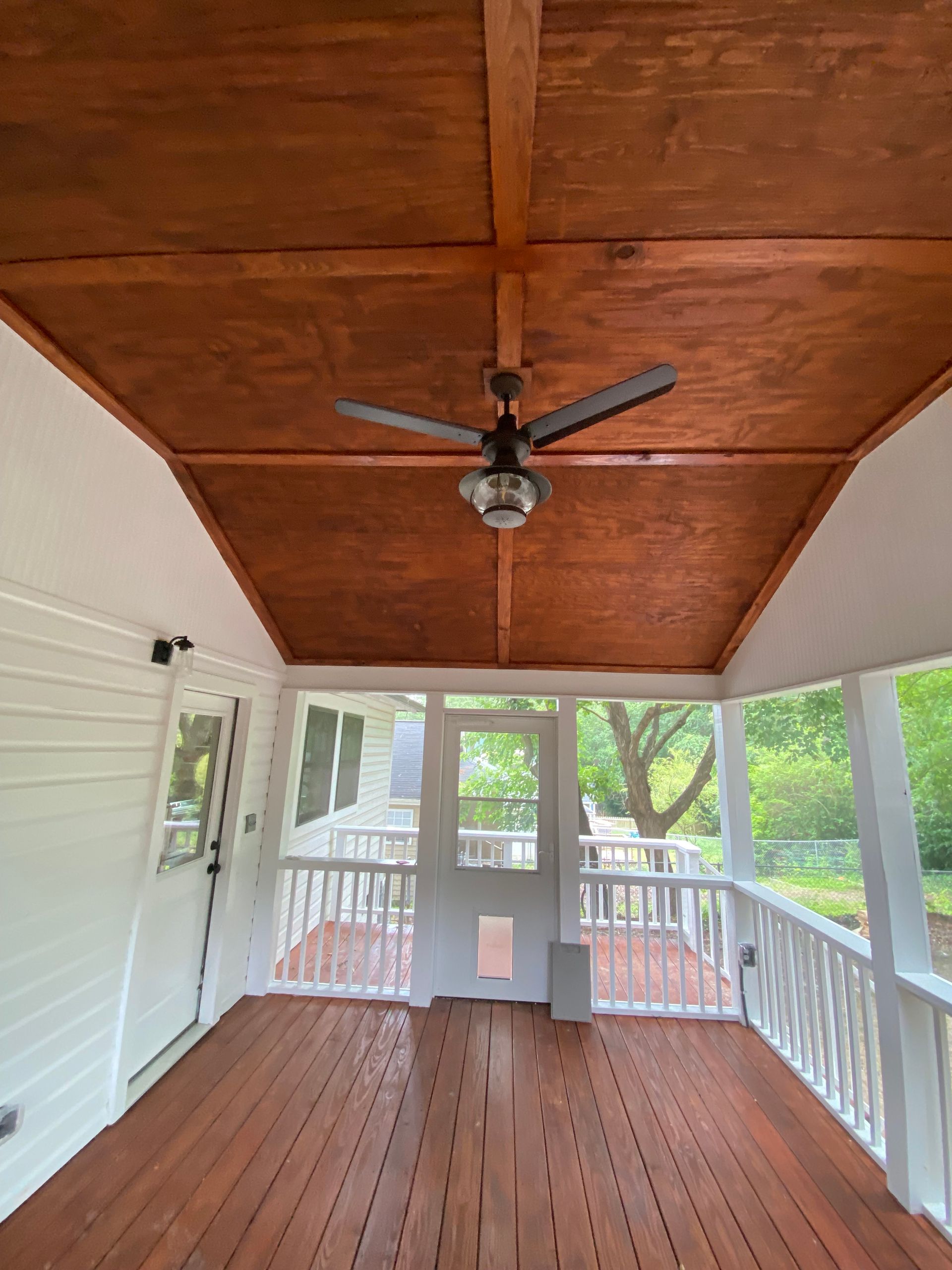 A screened porch with a stained wood ceiling, ceiling fan, and white railing, looking out onto a yard.