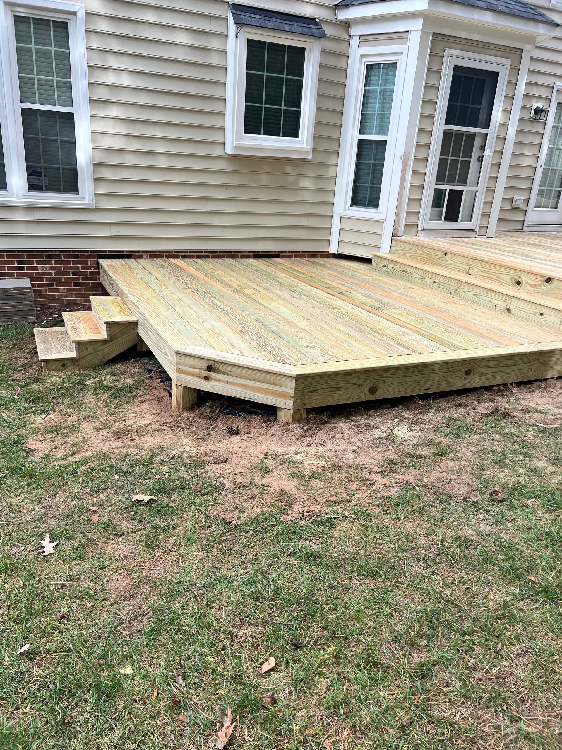 Wooden deck with steps attached to a tan house. The deck is unfinished, sitting on grass.