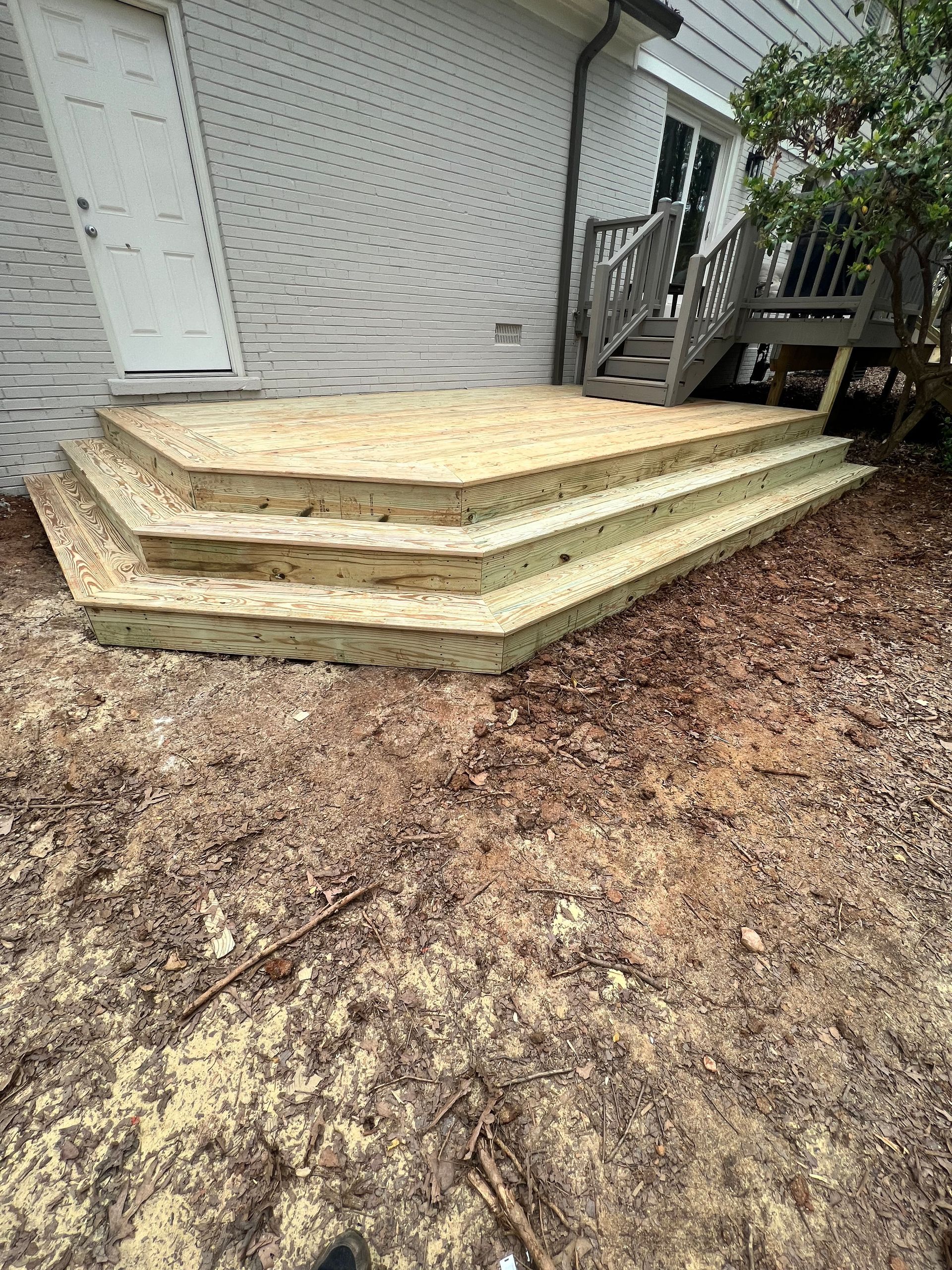 Wooden deck with steps against a gray house, next to a door. Ground is dirt.