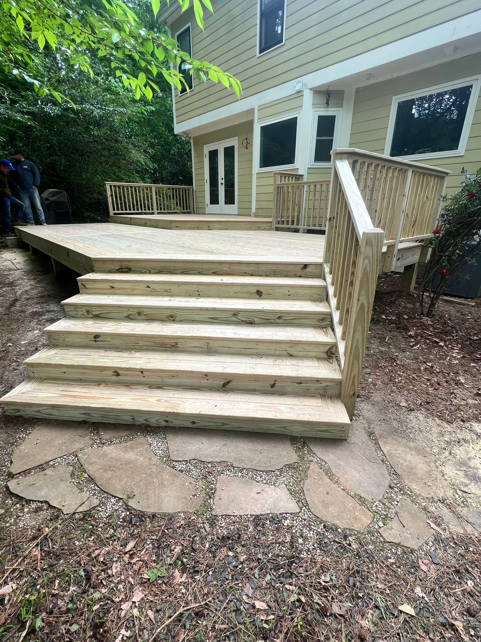 Wooden deck with stairs leading down to a stone path. House in the background, surrounded by trees.