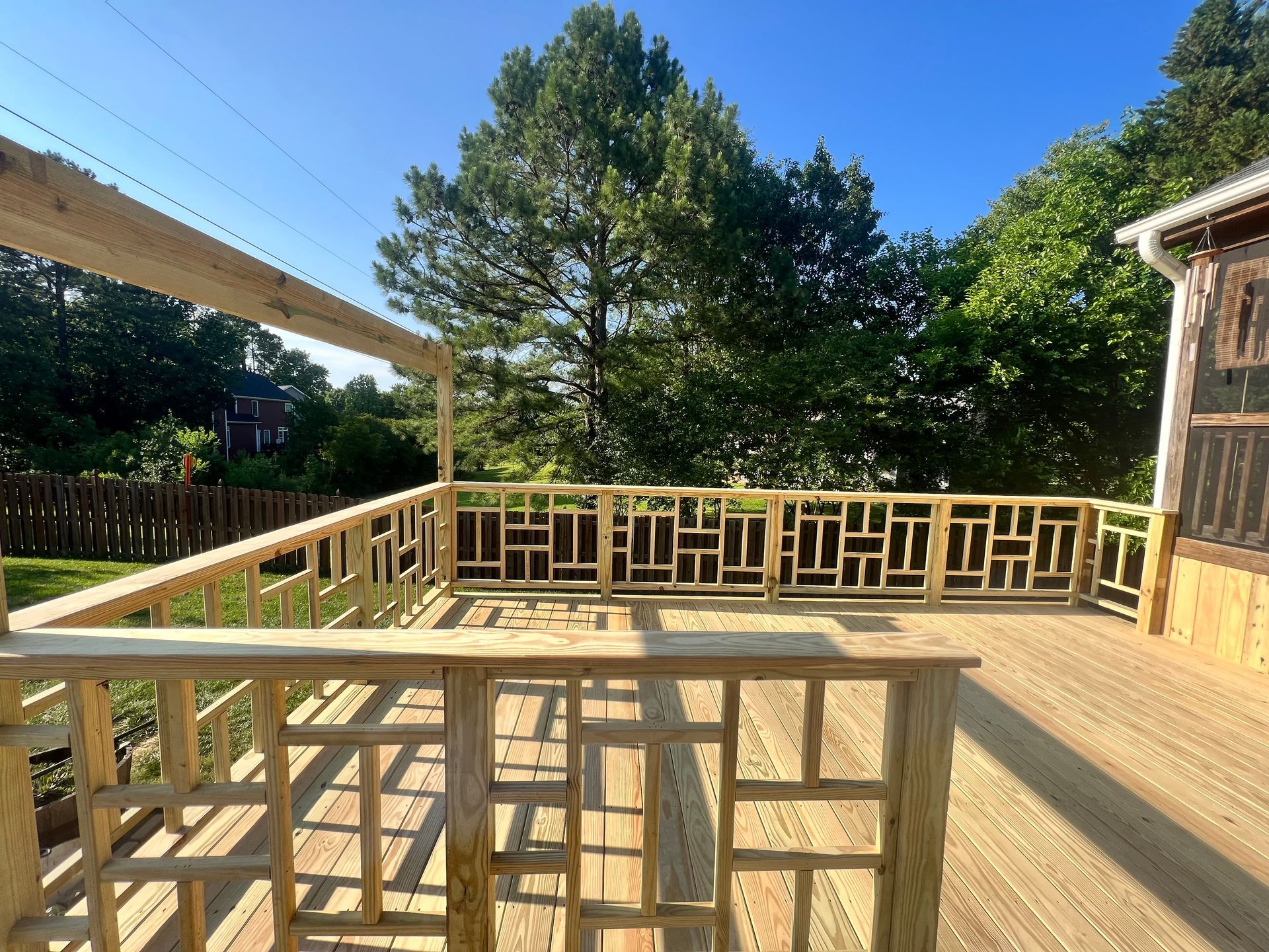 Wooden deck with decorative railing, surrounded by green trees under a blue sky.