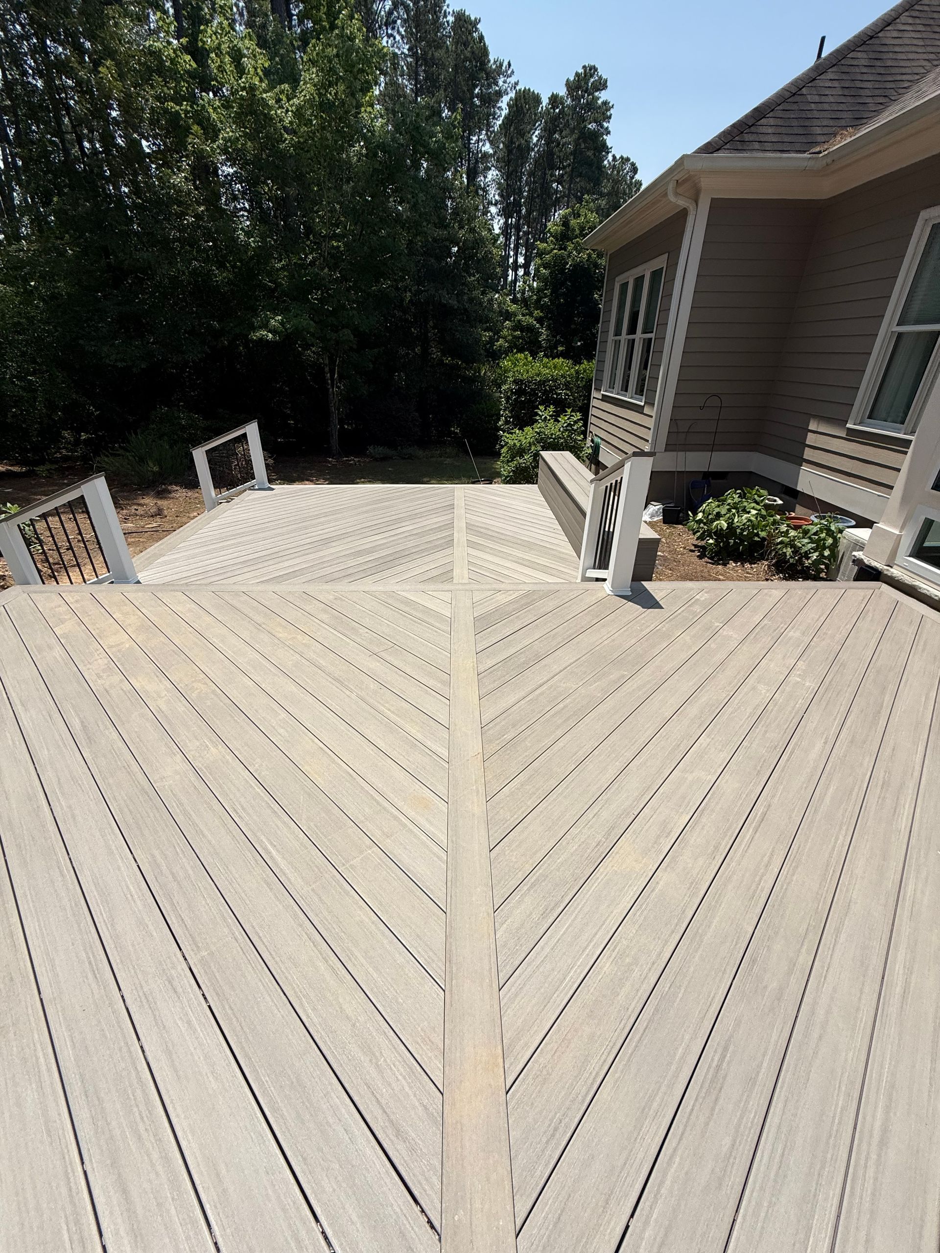 Angled view of a wooden deck with a patterned design; next to a house with lush green trees and a bright blue sky.