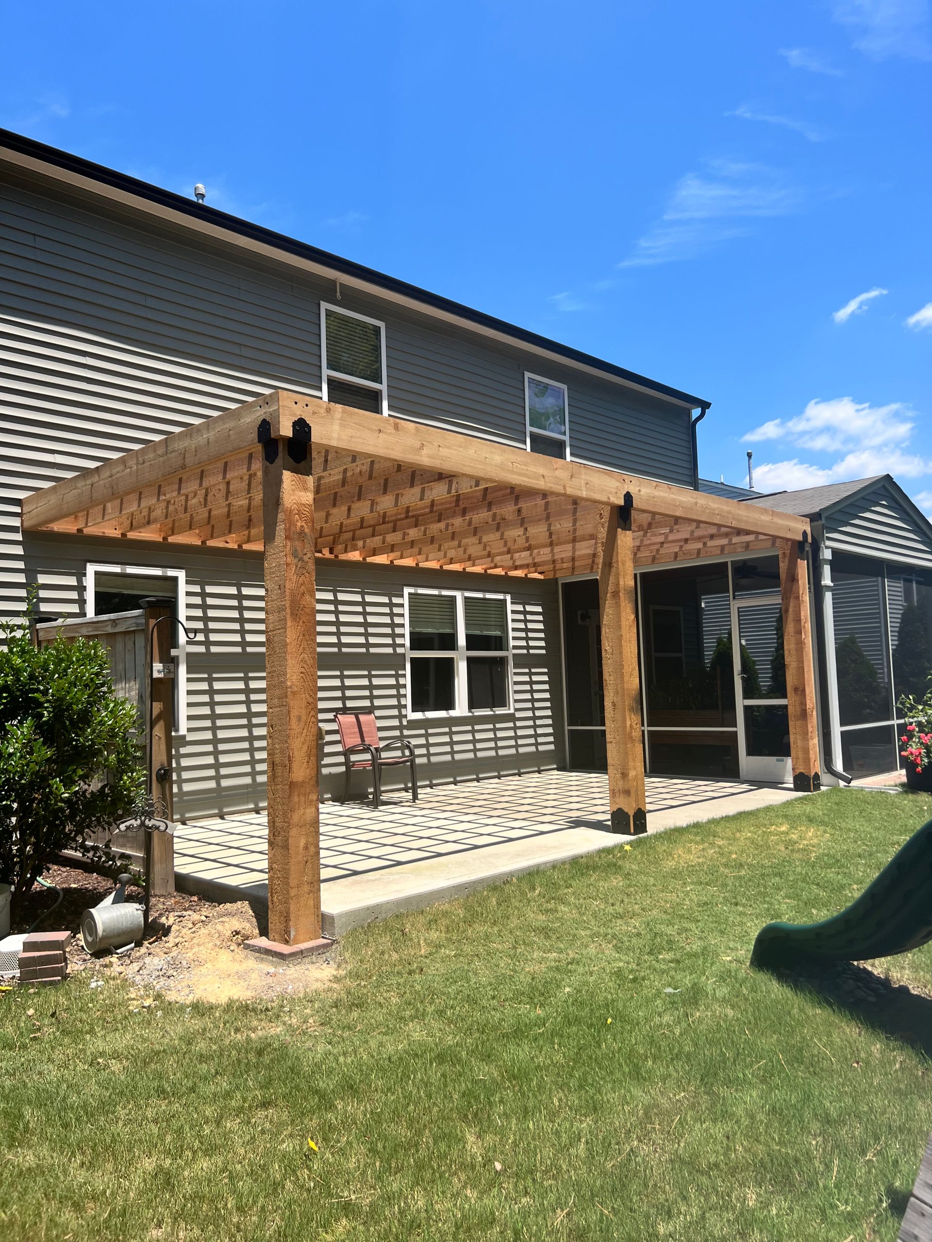 Wooden pergola attached to a gray house with a concrete patio and green lawn on a sunny day.