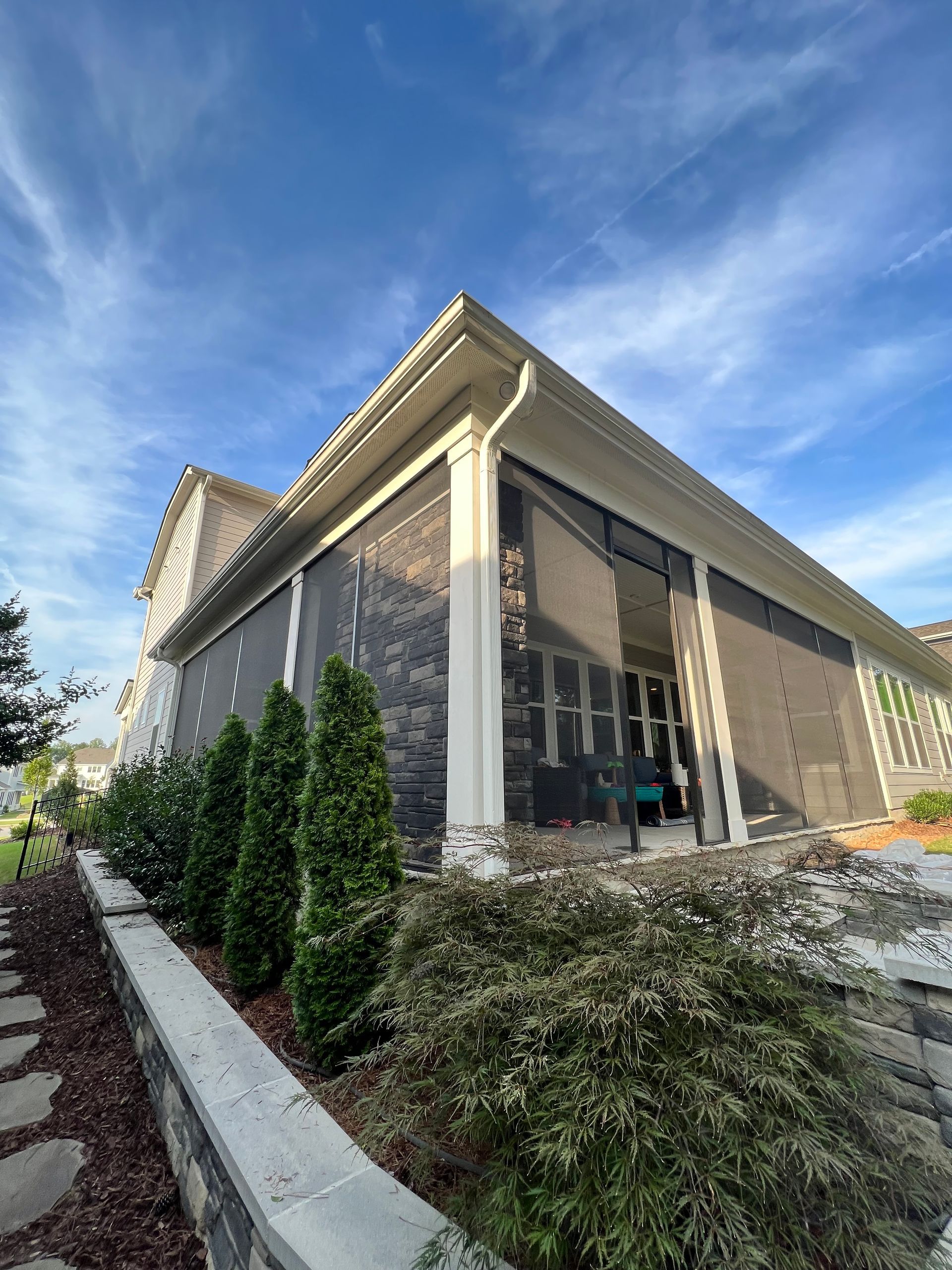 A screen-enclosed porch attached to a light-colored house with landscaping under a blue sky.
