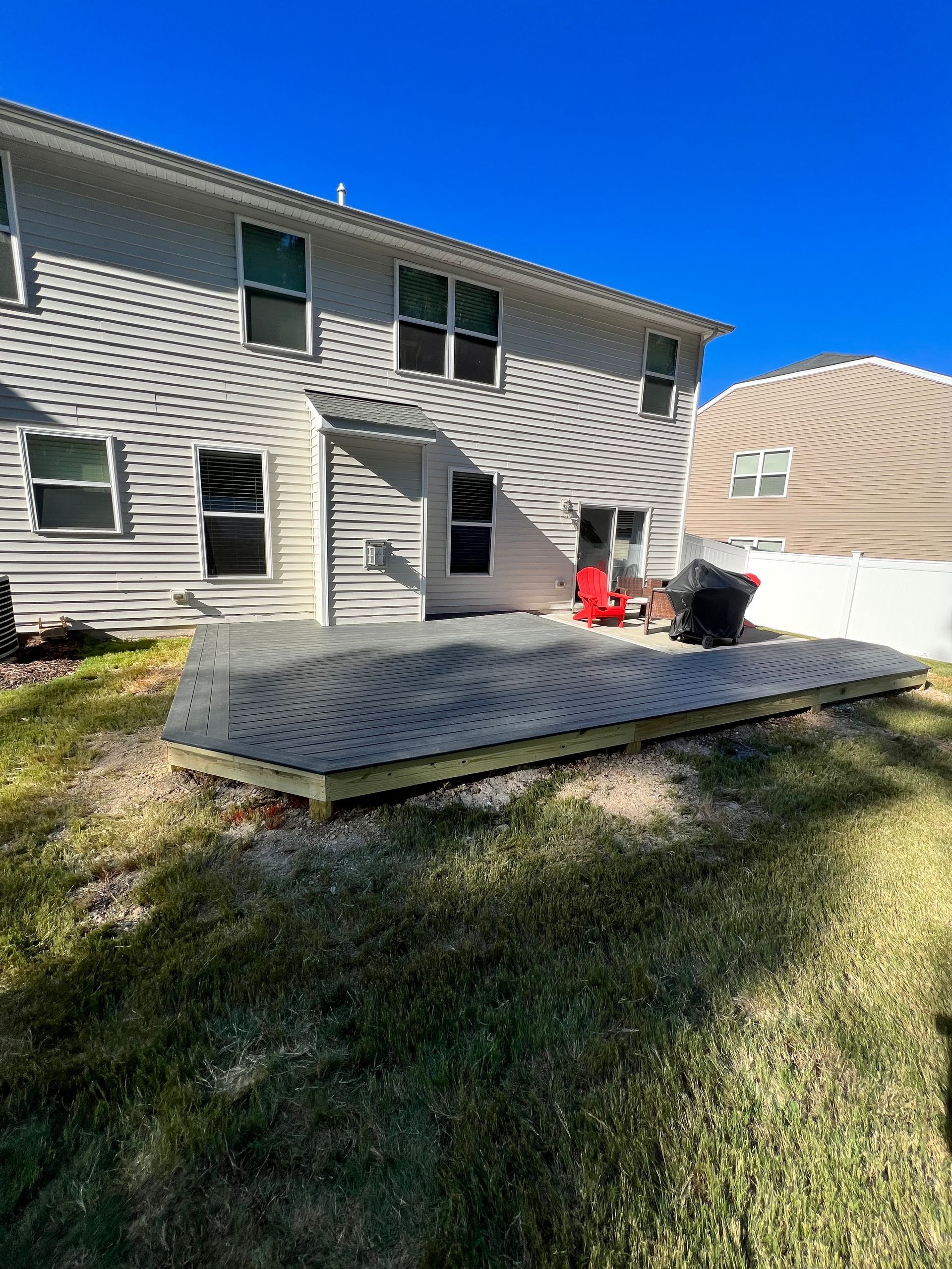 A gray deck extends from a two-story house, with a red chair and grill on the deck.